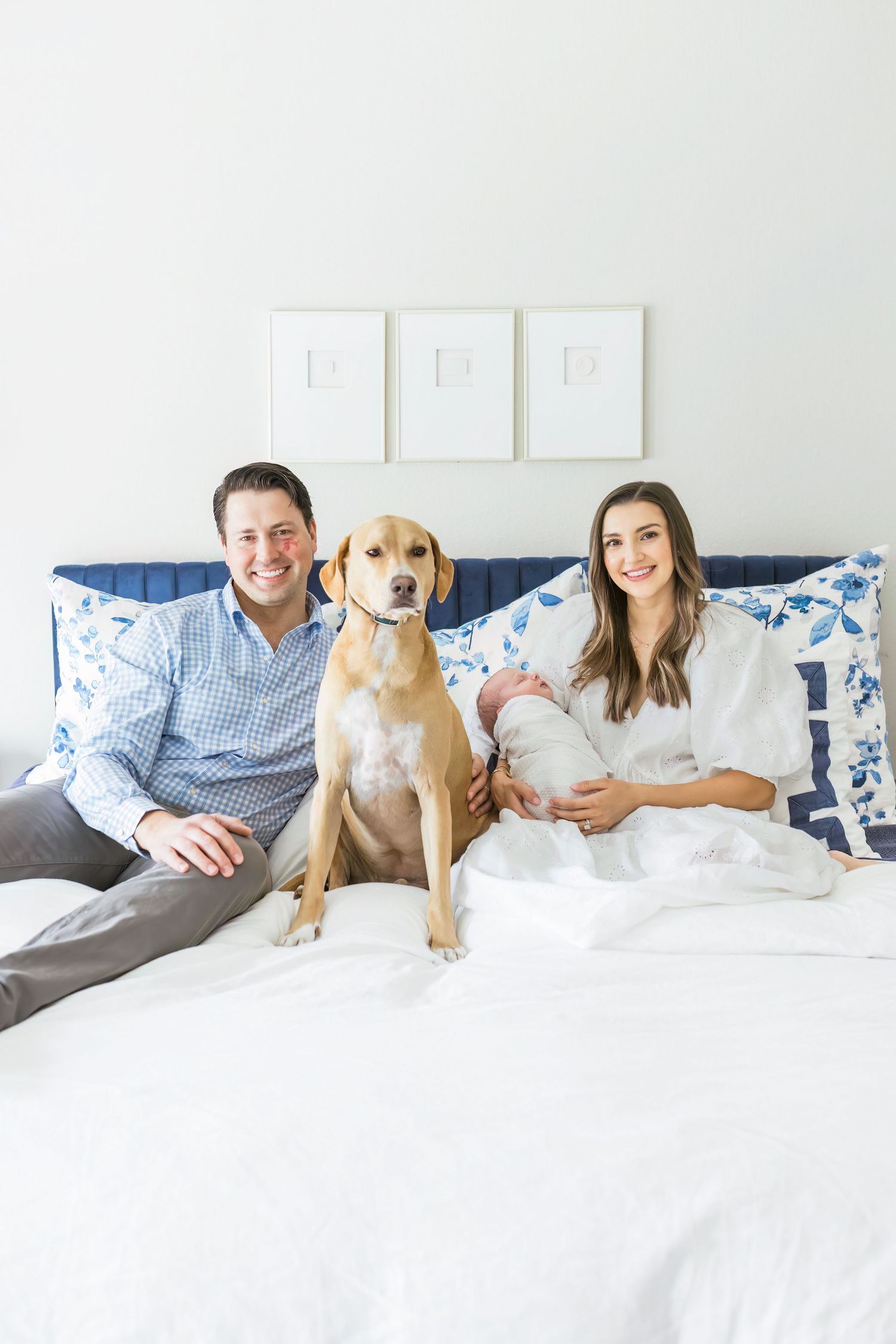 Family portrait on white bed with yellow lab dog and blue decorative pillows.