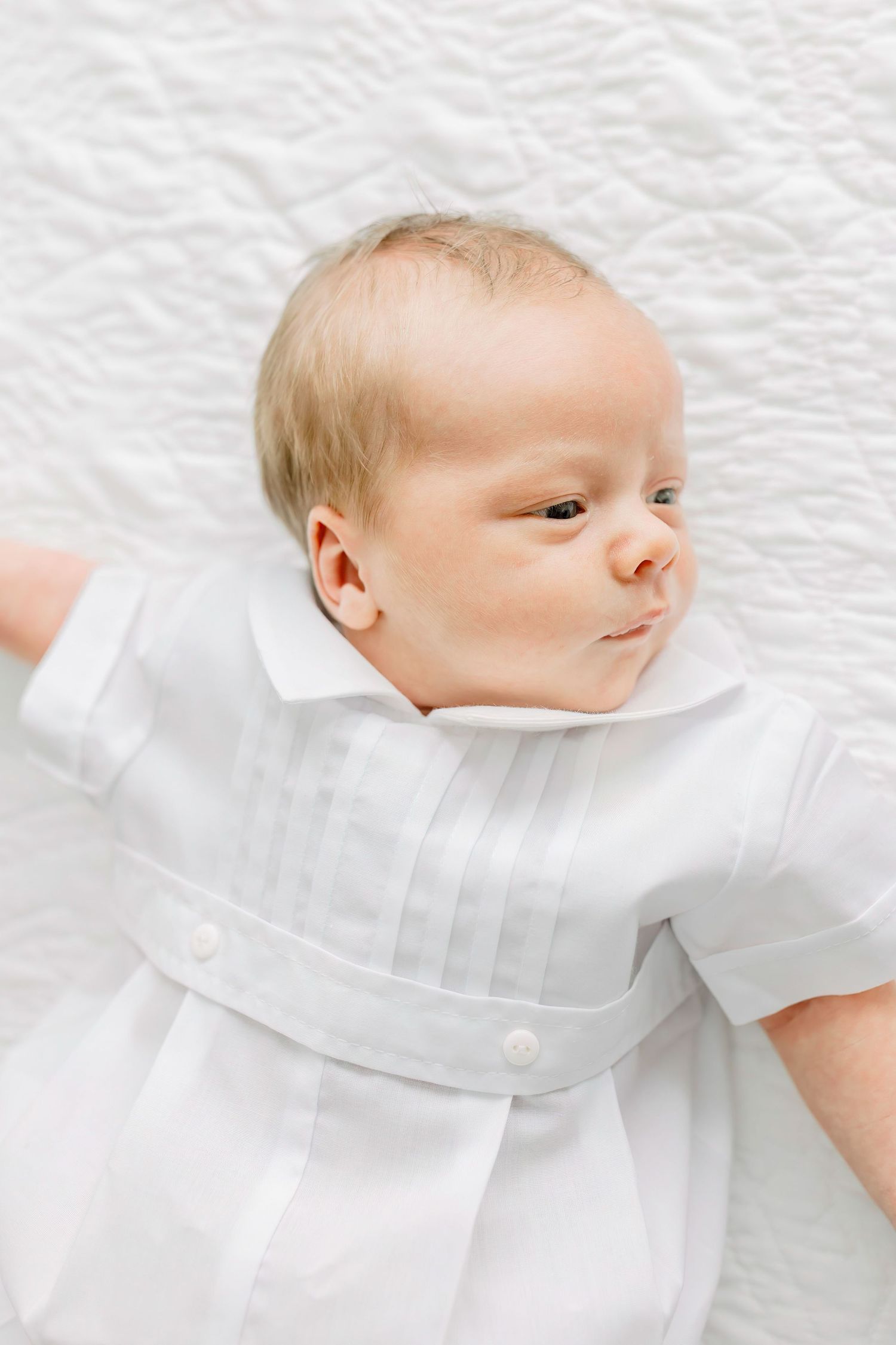 Baby in white christening gown lying on textured white fabric.