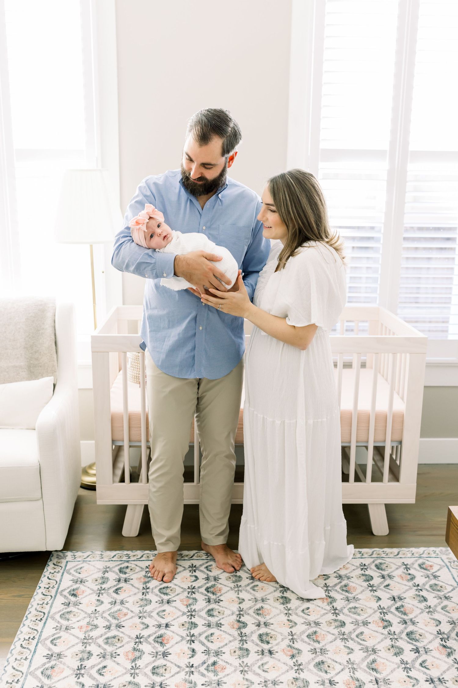 A couple in blue and white outfits stands lovingly with their newborn baby in a nursery with a patterned rug.