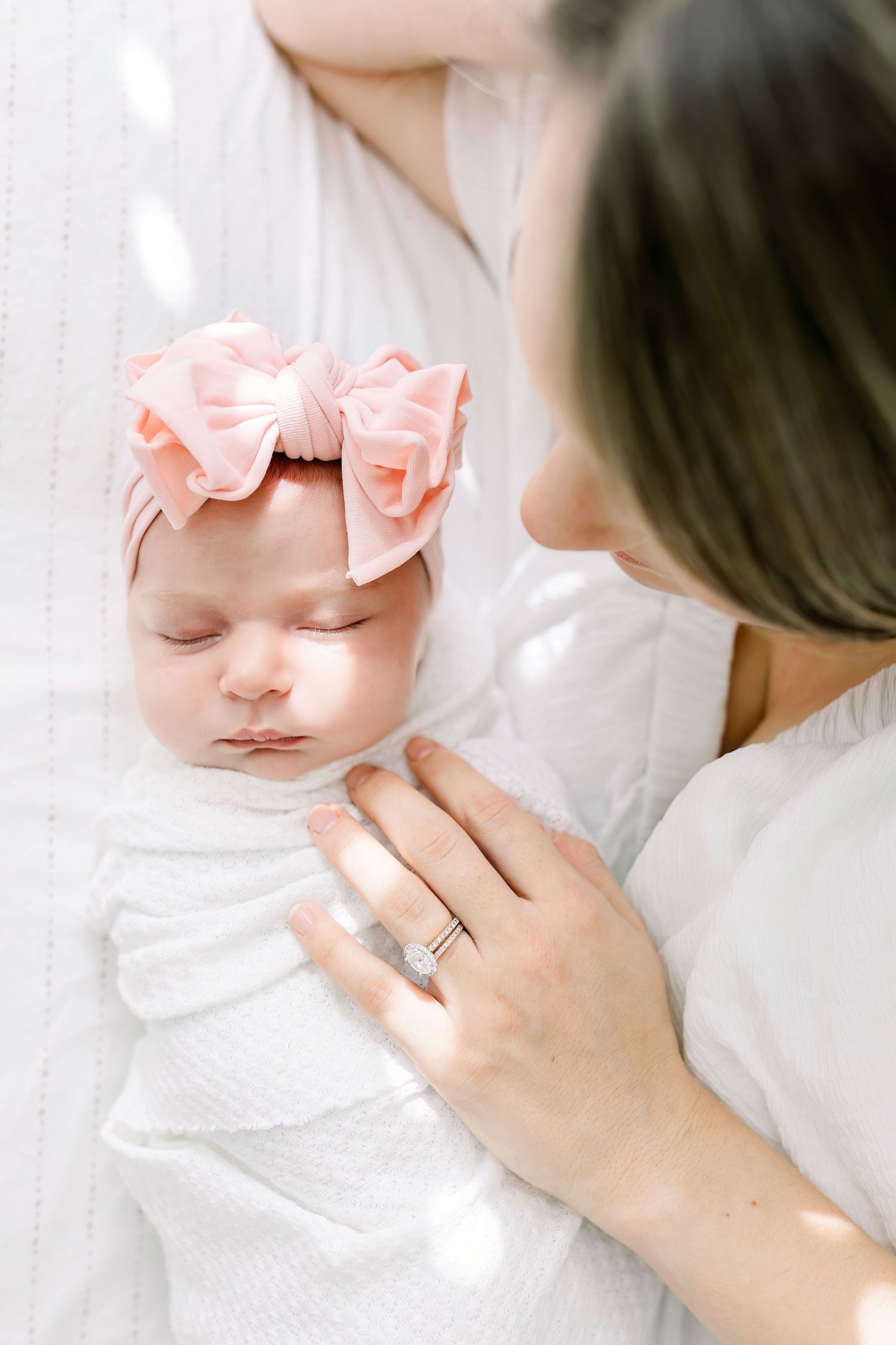 Sleeping newborn baby wearing a pink bow headband rests peacefully on white bedding.