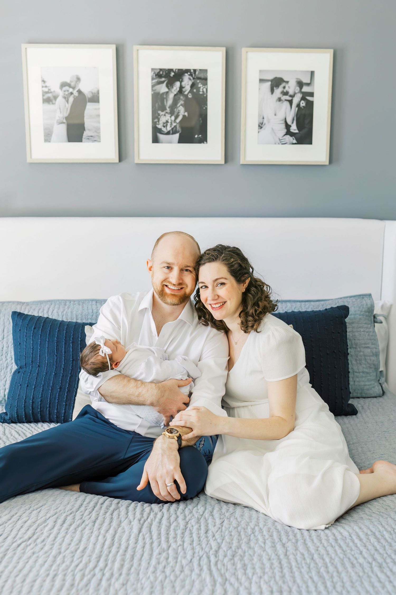 A couple in white outfits sits on a gray bed with throw pillows and framed photos on the wall.