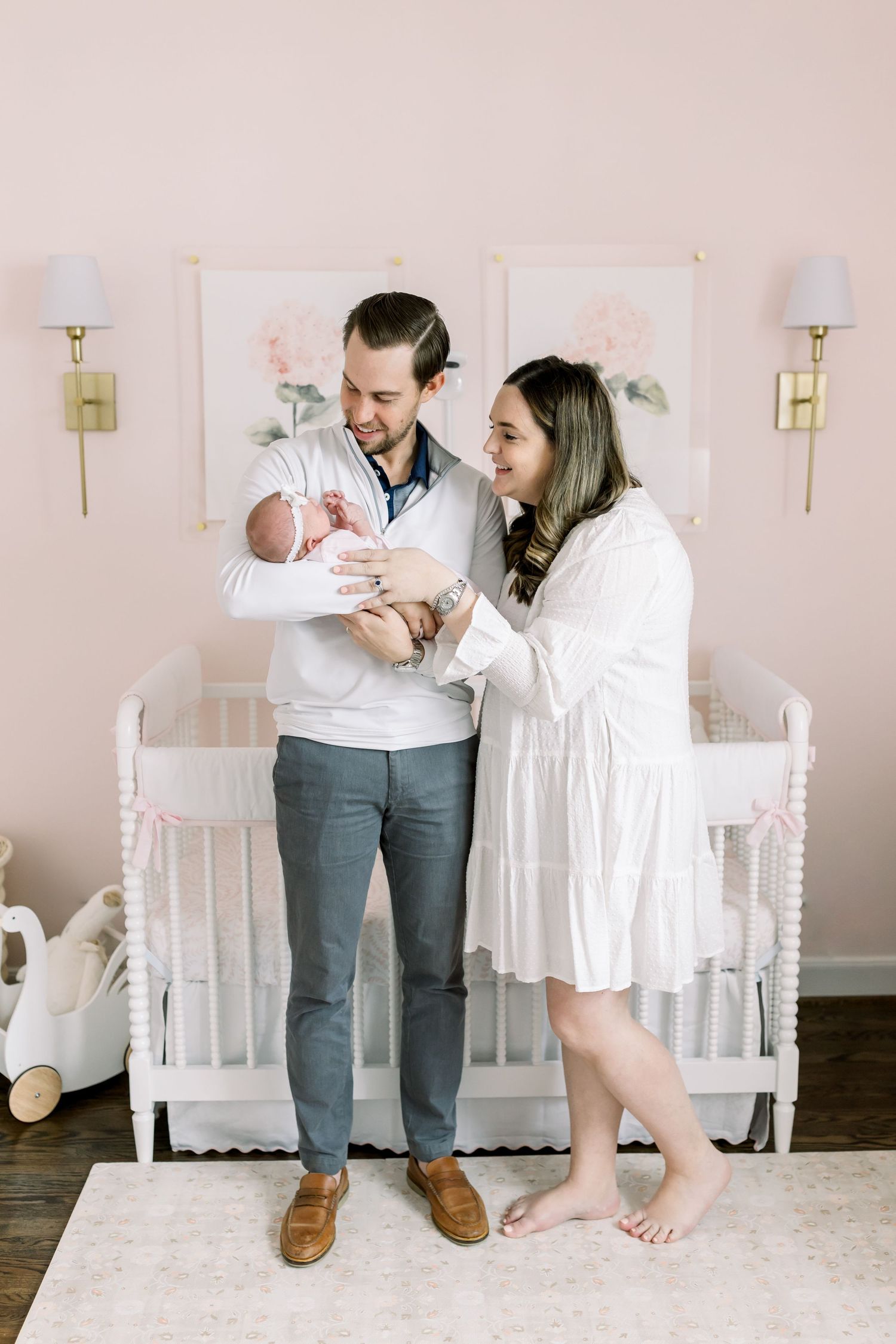 A happy couple stands in a pink nursery looking lovingly at their newborn baby wrapped in white.