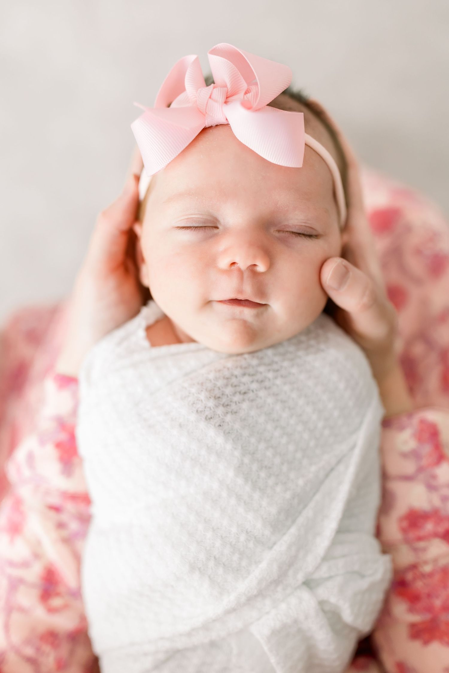 Peaceful sleeping newborn wrapped in white with pink bow headband.