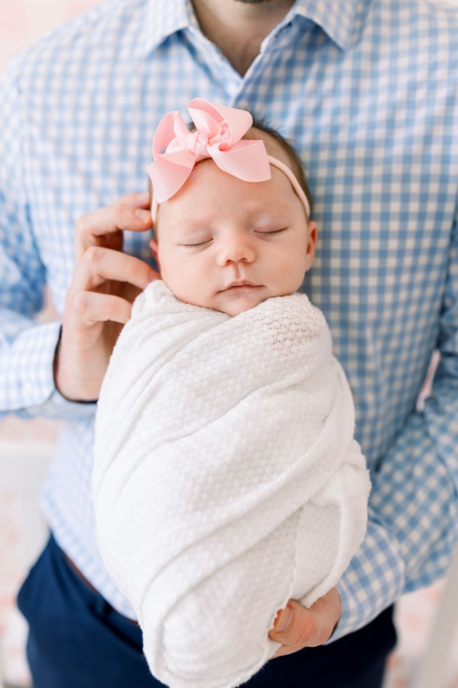 A sleeping newborn baby wearing a pink bow headband is swaddled in white while being held against a blue gingham shirt.