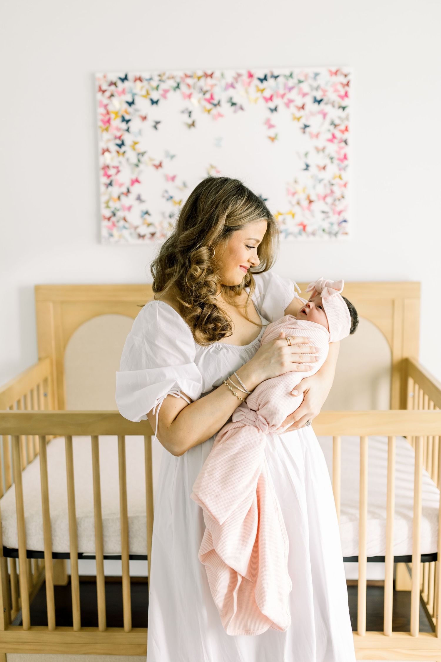Tender nursery moment in white dress beside wooden crib with colorful butterfly wall art.