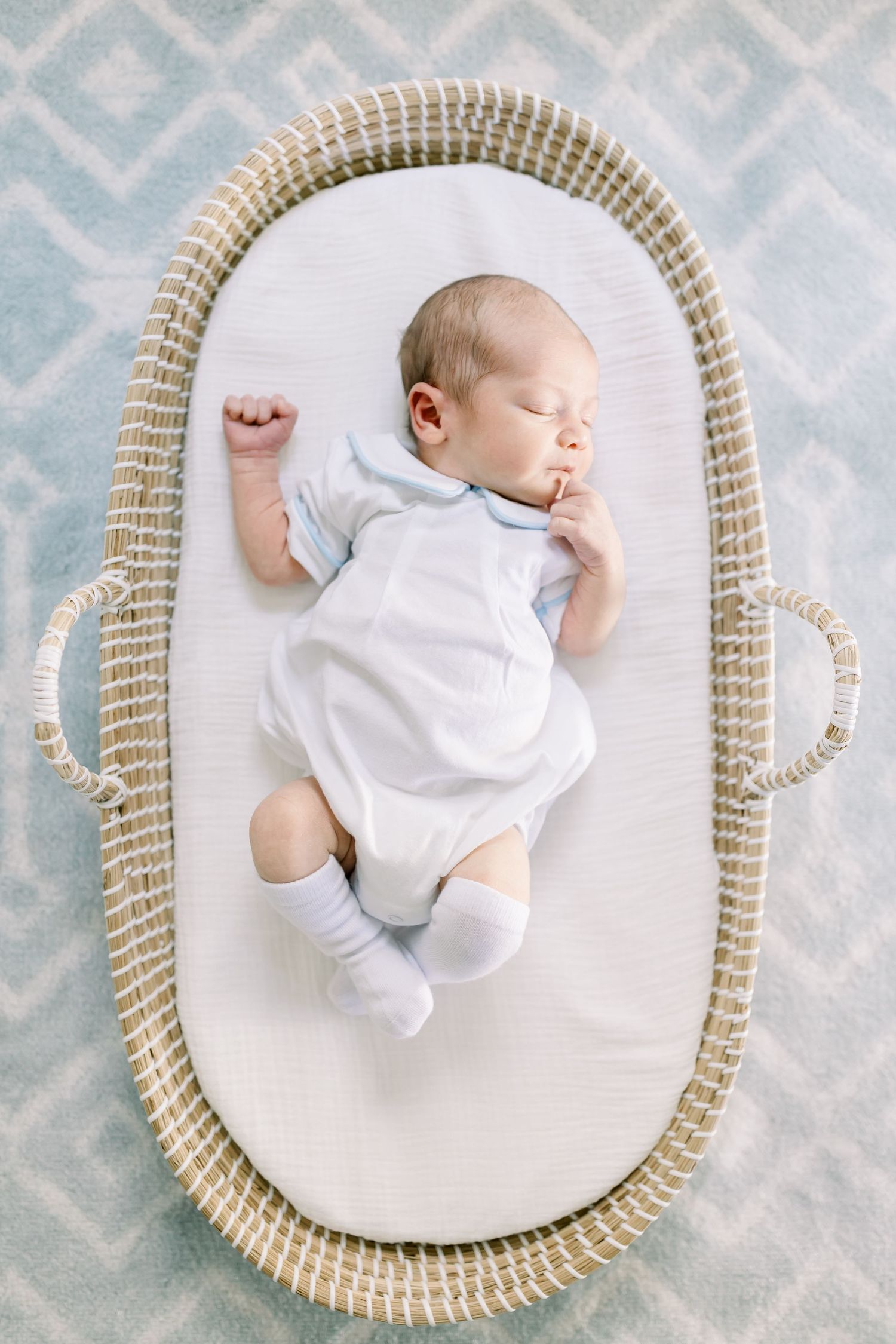 Sleeping newborn in white onesie lying in woven moses basket on patterned rug.