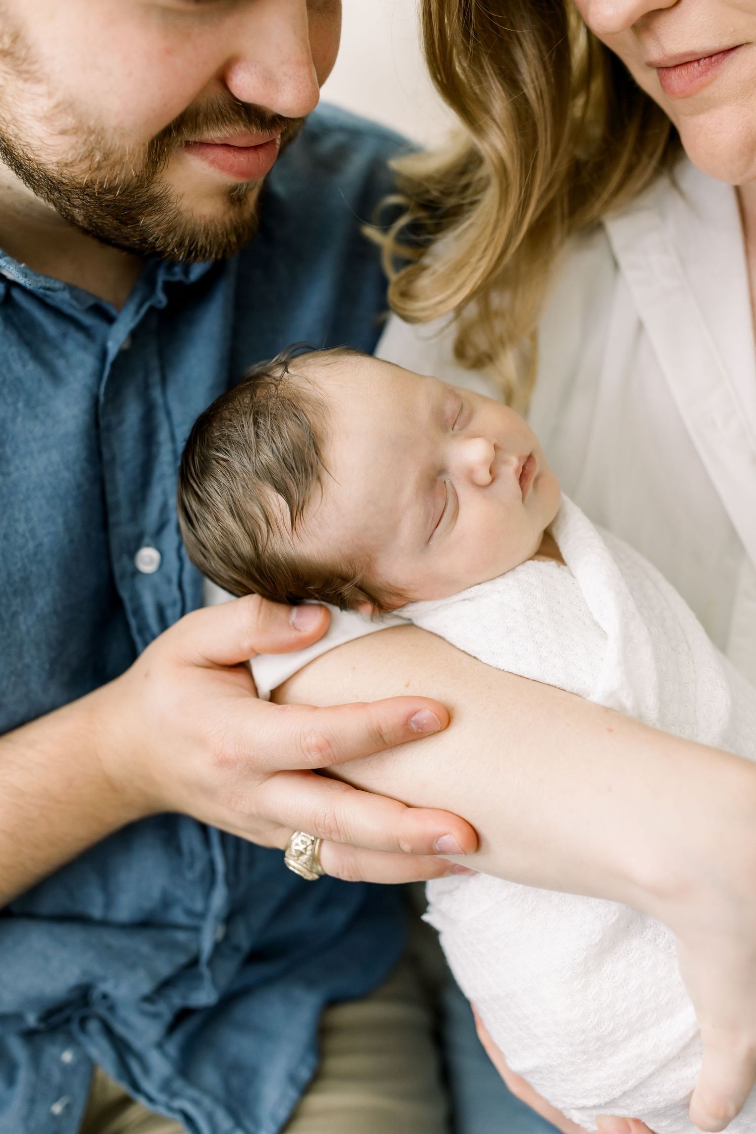 An intimate family moment as parents gaze down at their sleeping newborn swaddled in white.