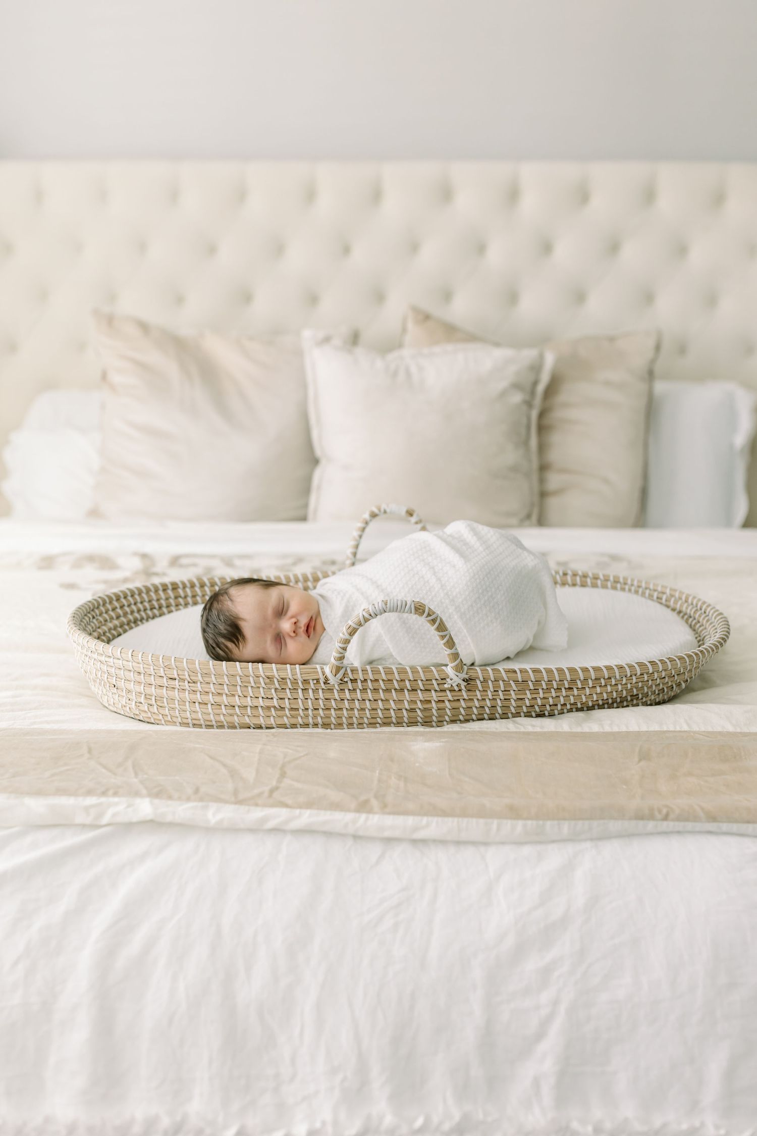 A sleeping newborn baby lies in a woven basket on a plush white bed.