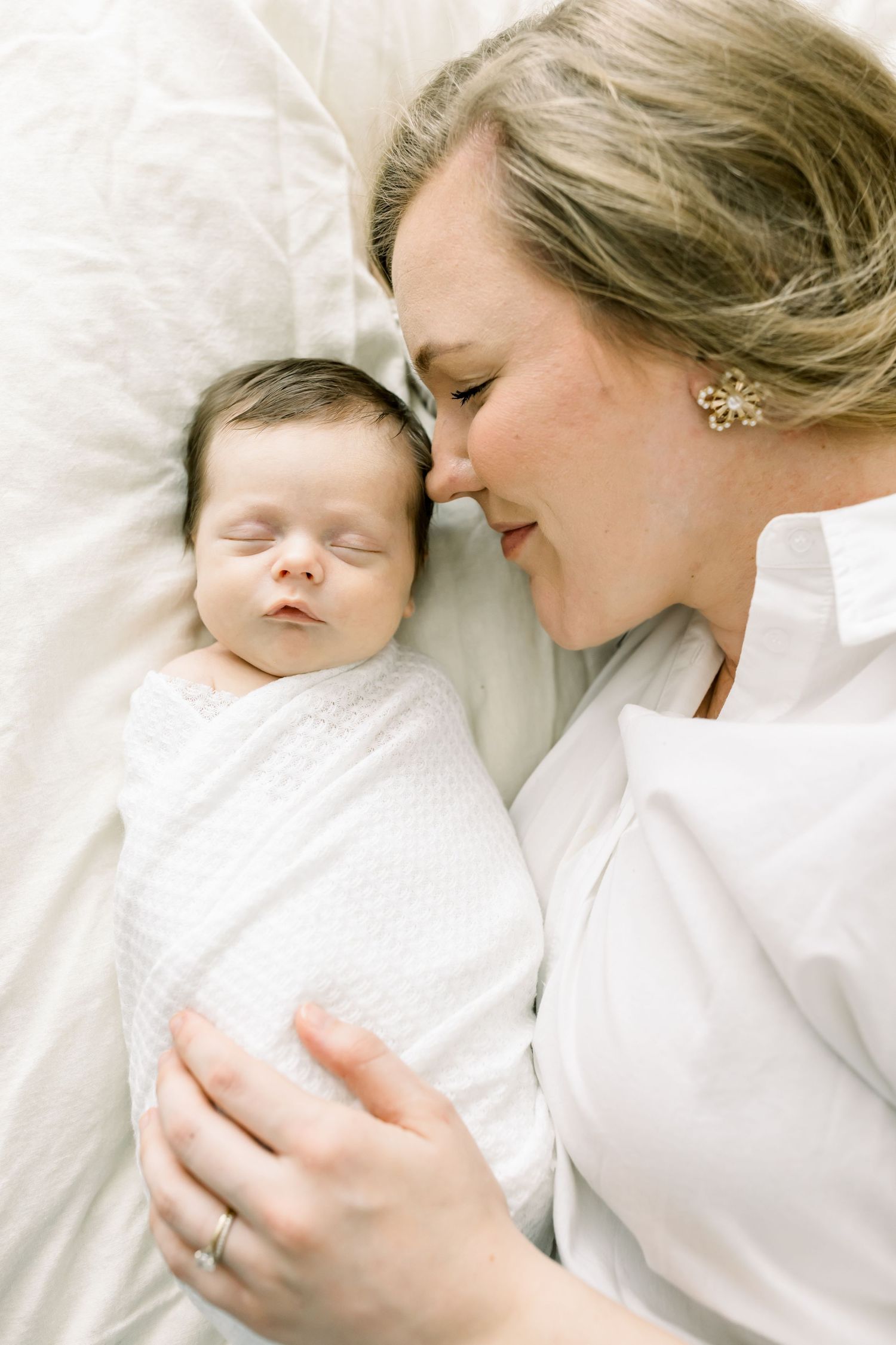 An intimate portrait of a tender moment between mother and sleeping baby on white bedding.