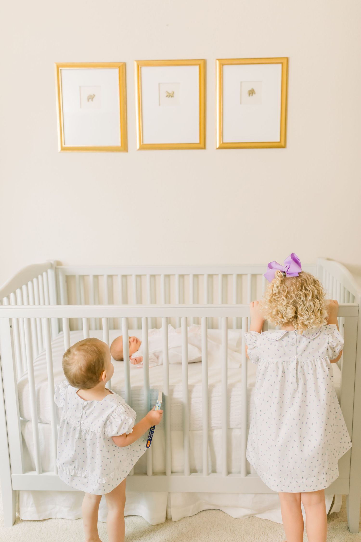 Two small children in white dresses looking into a white crib beneath framed artwork.
