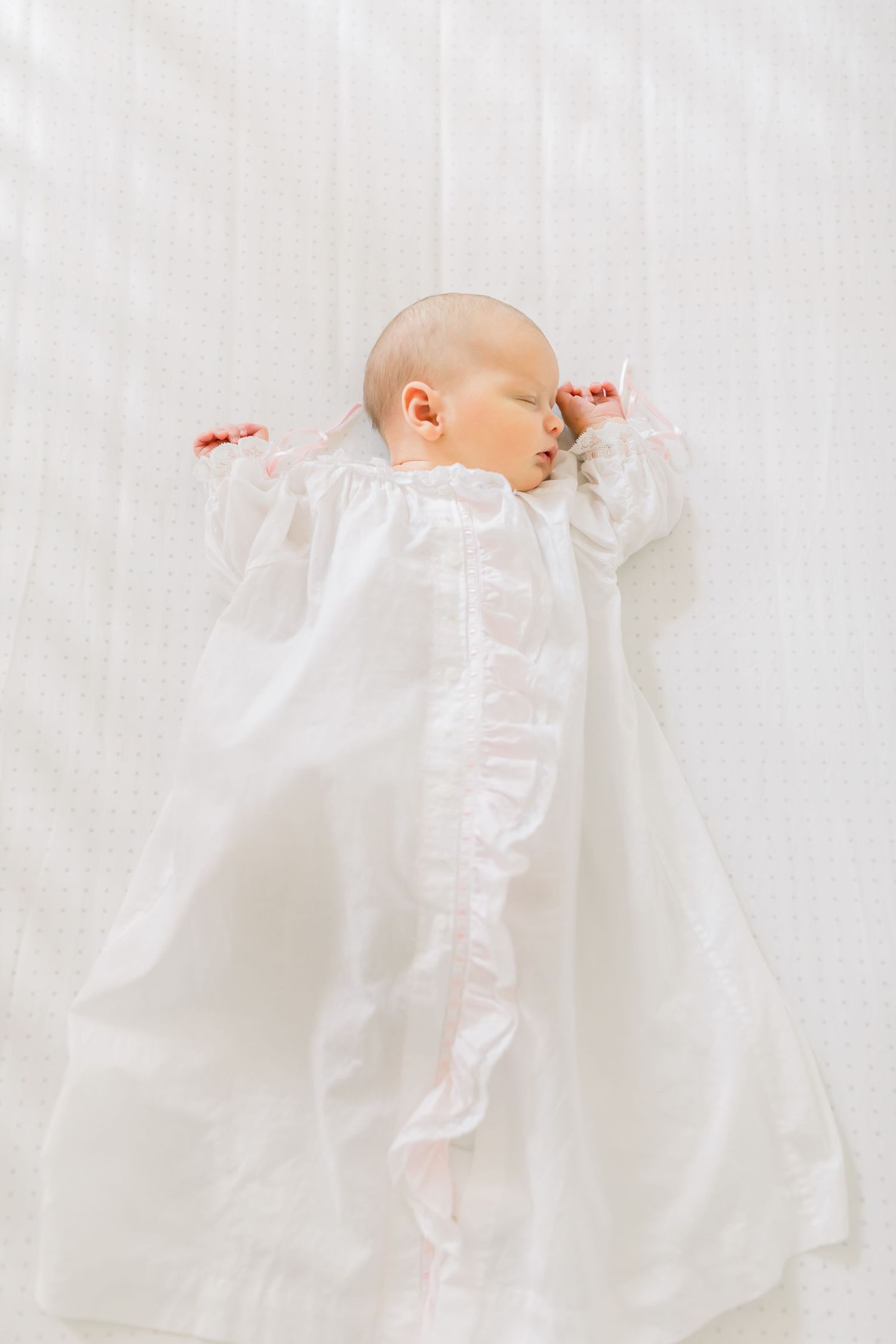 A sleeping baby wearing a white christening gown on light fabric.