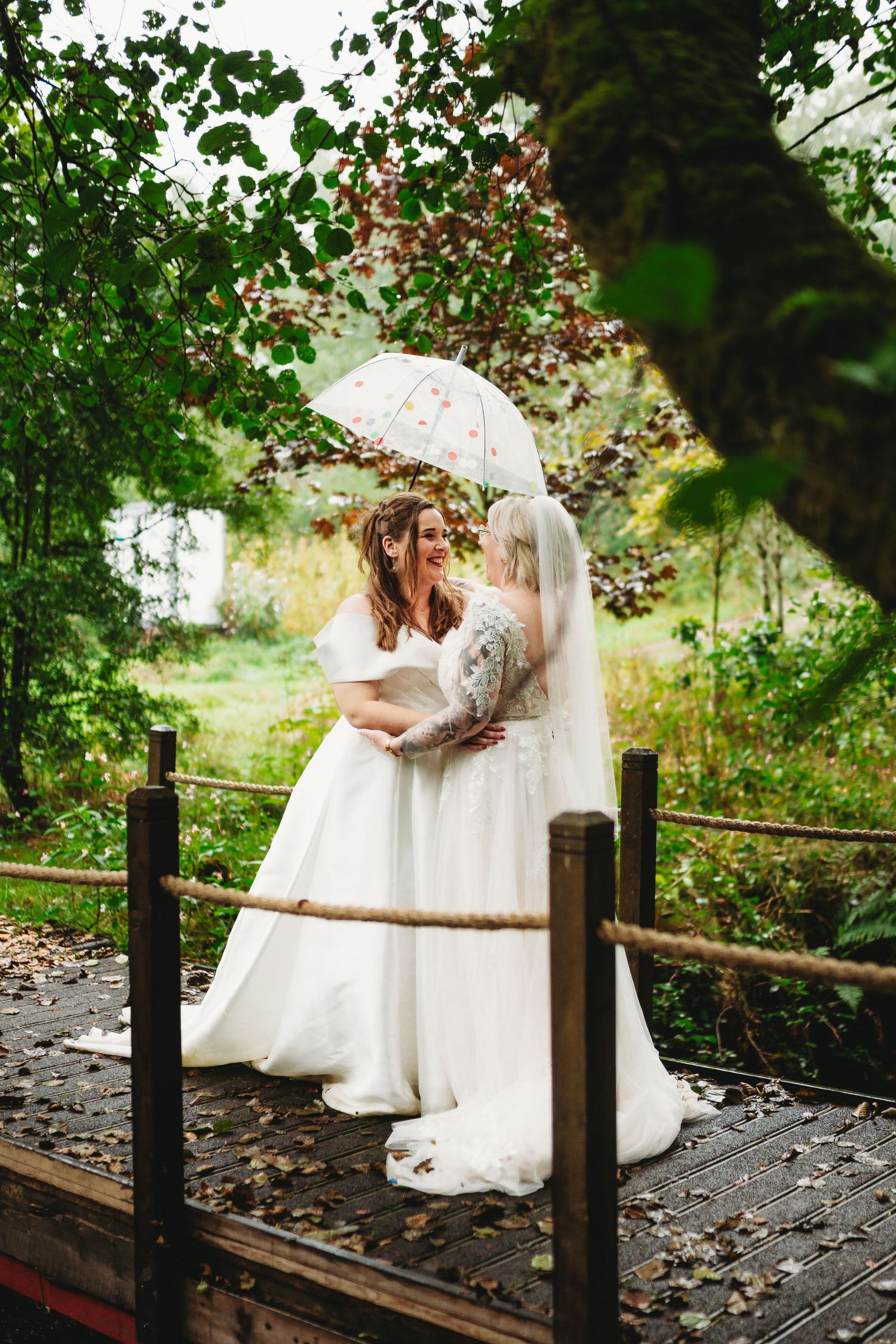 Two brides in white gowns embrace under anumbrella on a wooden bridge surrounded by lush greenery at Oak Tree Farm Weddings Carmarthan
