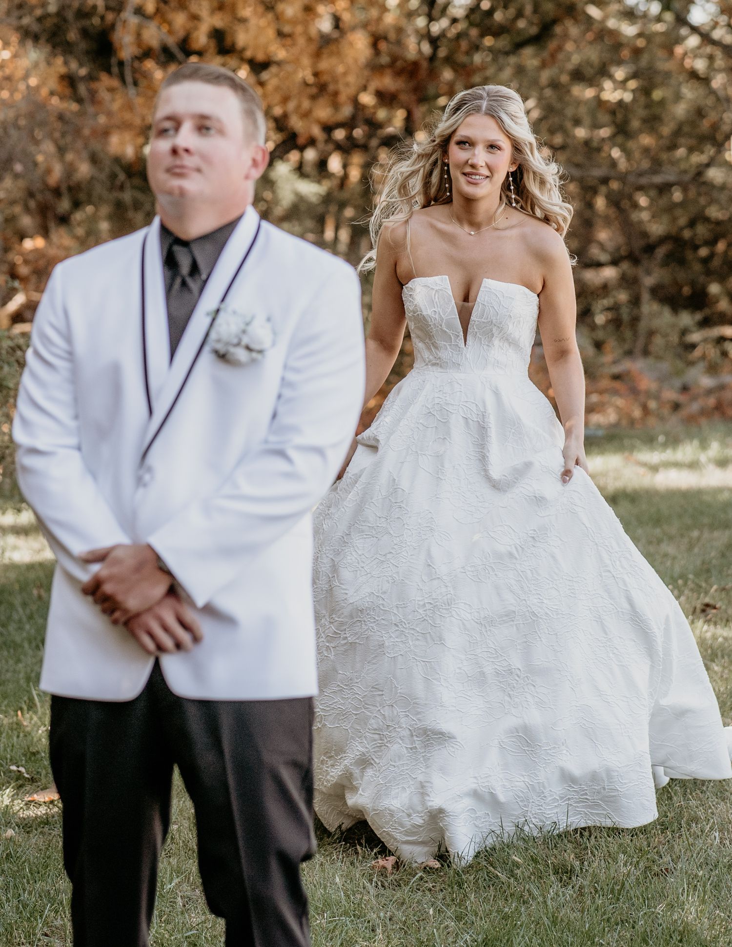 A bride in a strapless white gown poses with someone wearing a white tuxedo jacket during an autumn outdoor wedding shoot.