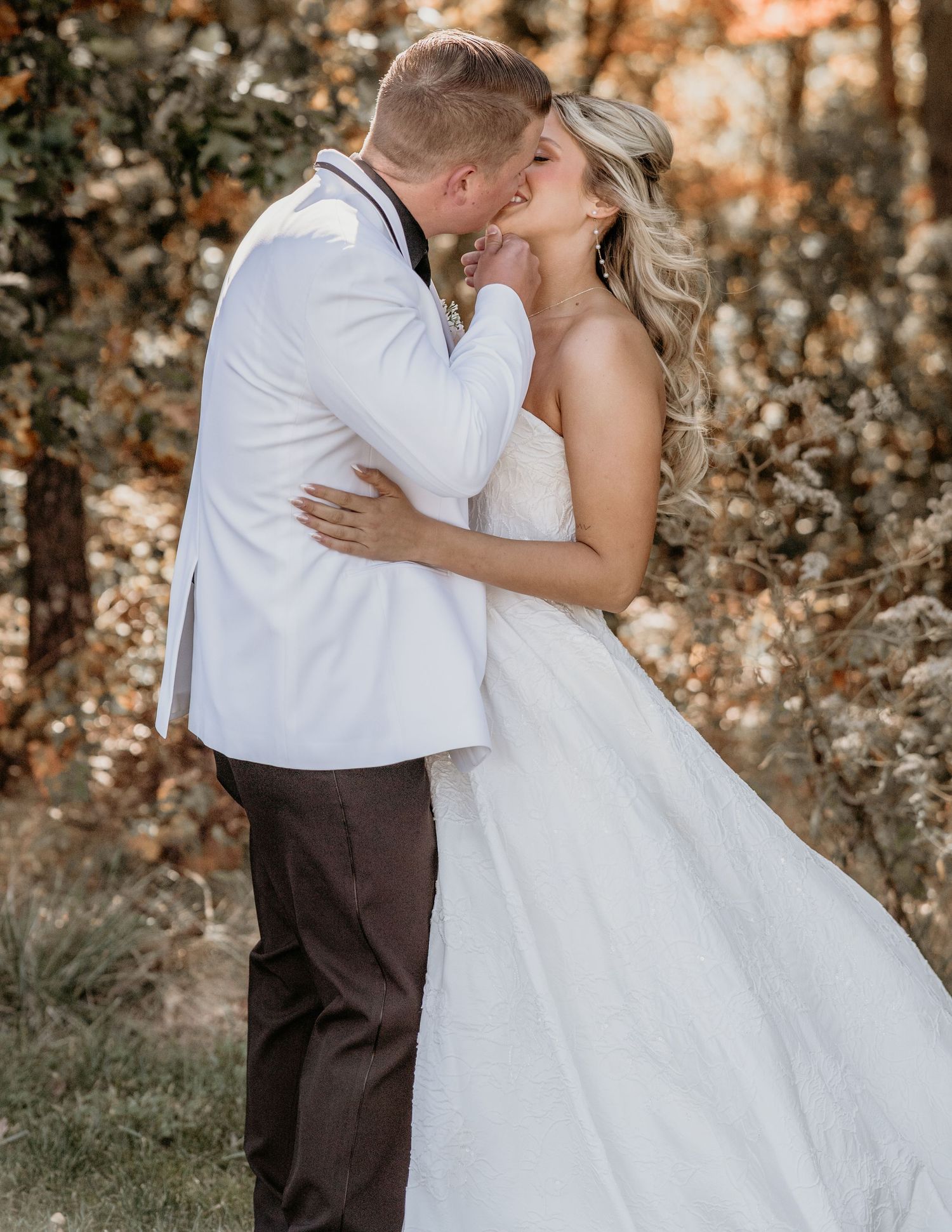 Bride and groom share a romantic kiss in an autumn forest setting with warm golden light filtering through the trees.