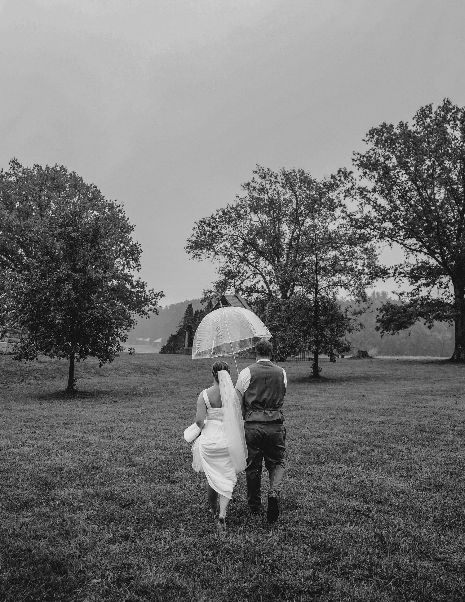 A romantic couple walking together under a white umbrella on a grassy field with a barn in the distance.