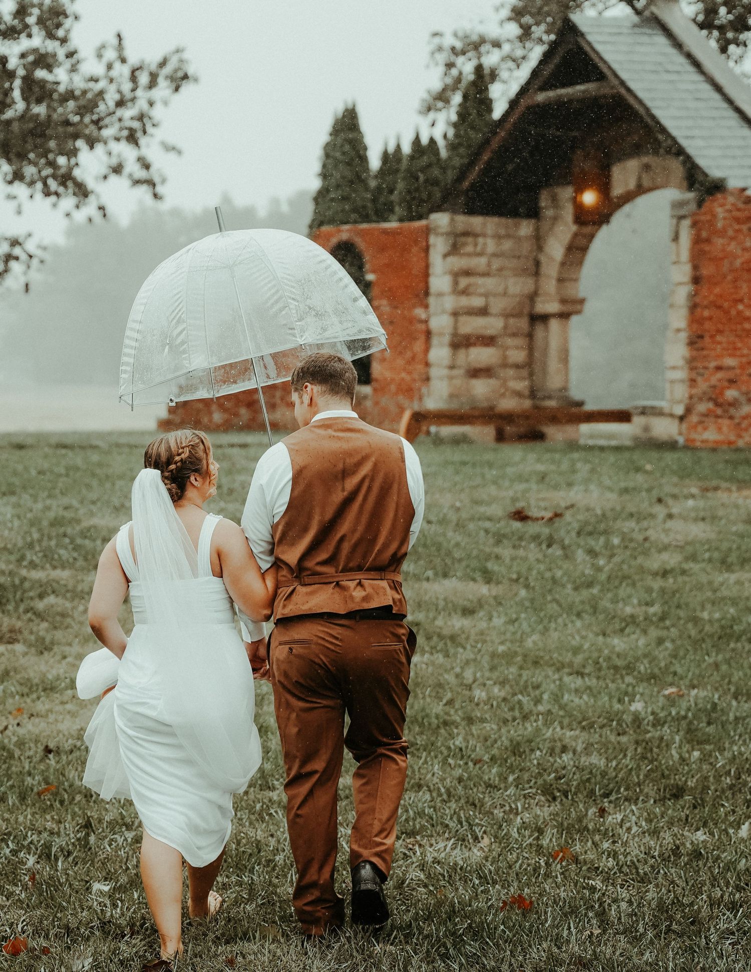 A couple sharing an umbrella as they walk in front of a misty stone archway on an overcast day.
