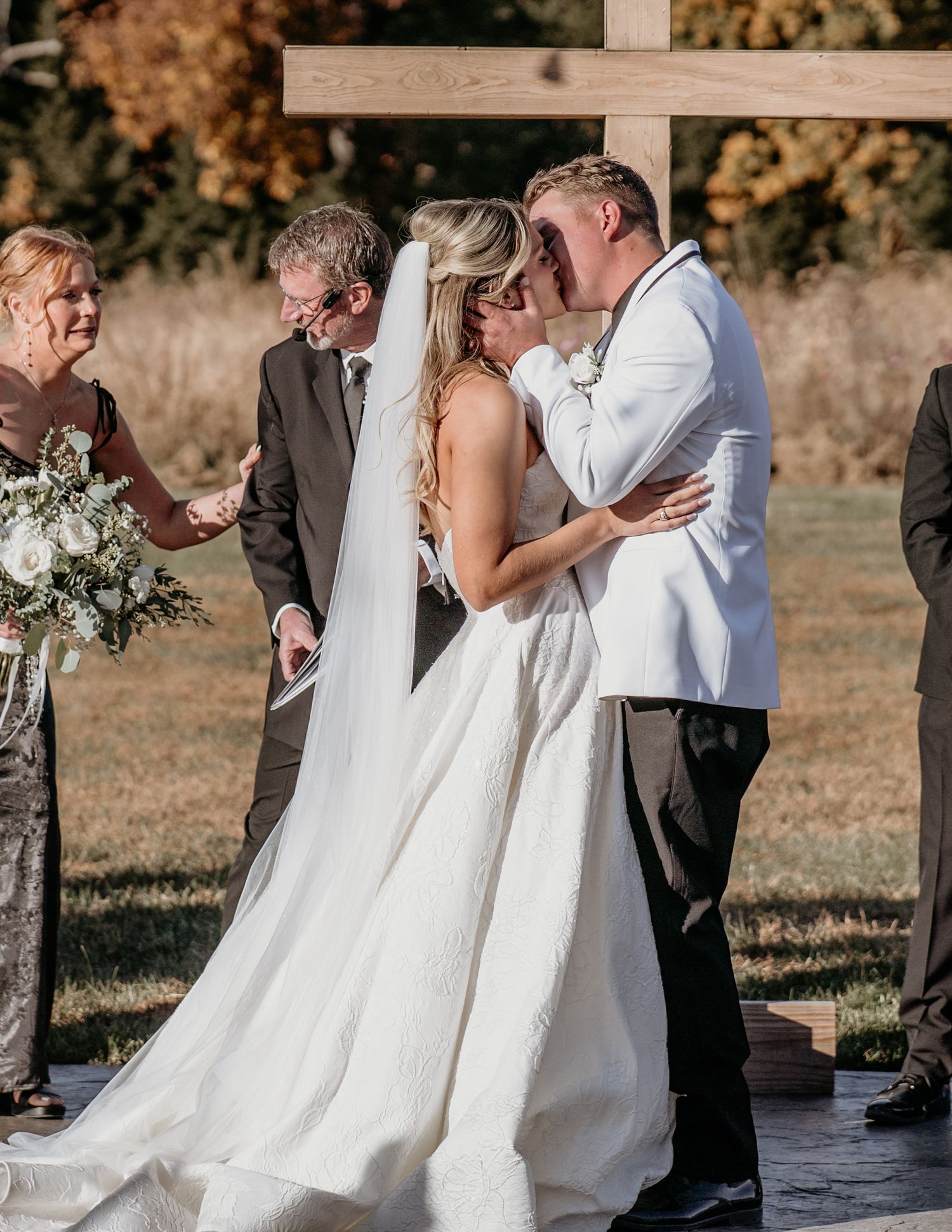 A wedding ceremony taking place outdoors with a wooden cross and autumn foliage in the background.