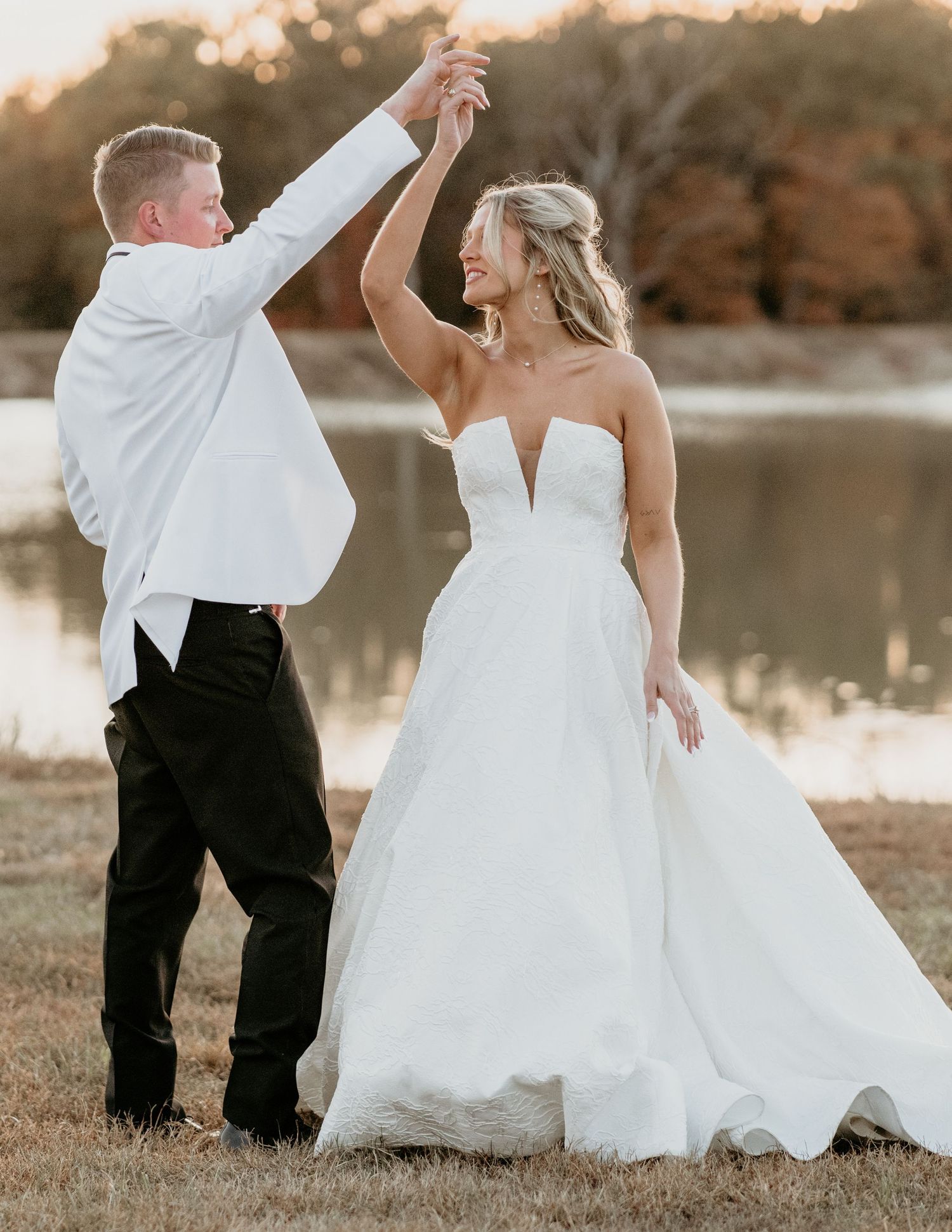 A bride in a white strapless gown dances with partner by a lake at sunset during their wedding celebration.