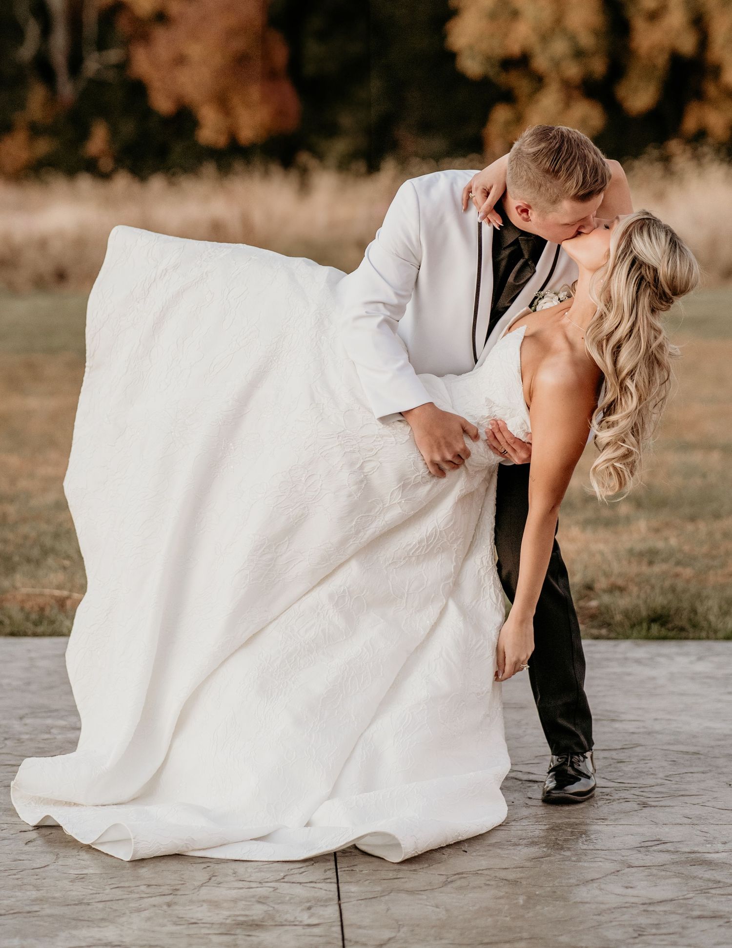 A romantic wedding photo showing a couple in elegant white dress and black suit dancing on a concrete patio at sunset.