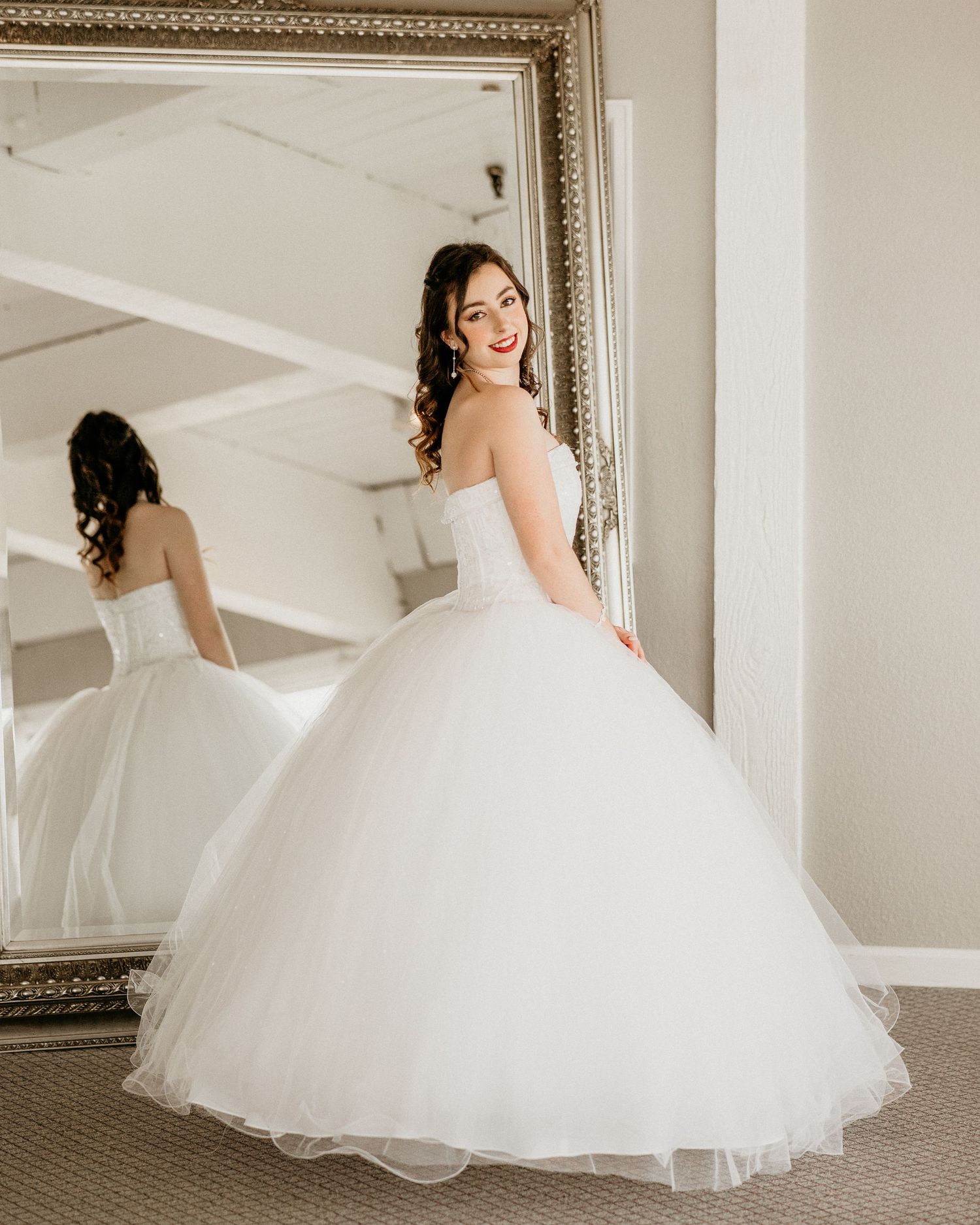A bride in a voluminous white tulle ball gown poses next to an ornate mirror in a bridal fitting room.