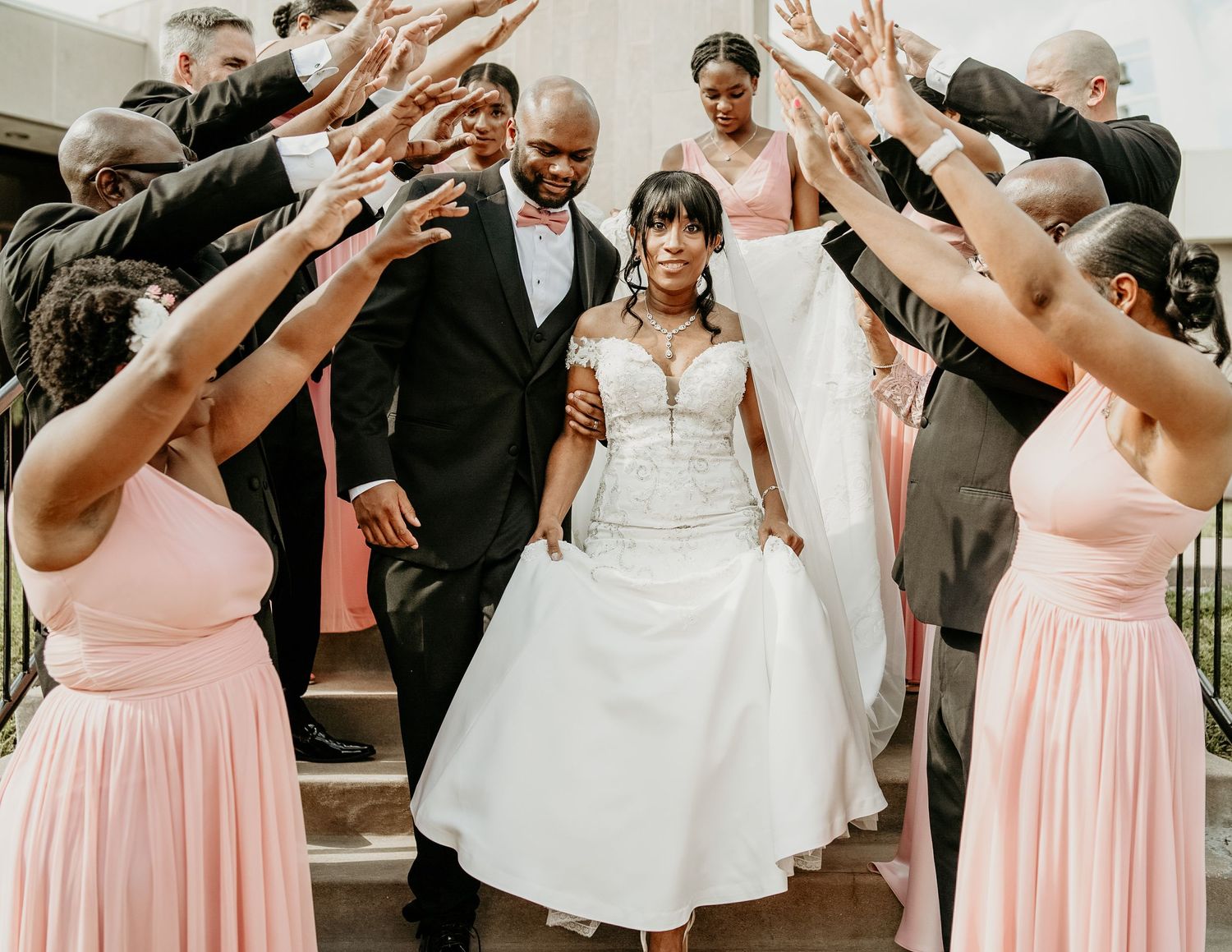 Wedding party in black suits and soft pink bridesmaid dresses forms a tunnel exit for the newlyweds on stone steps.