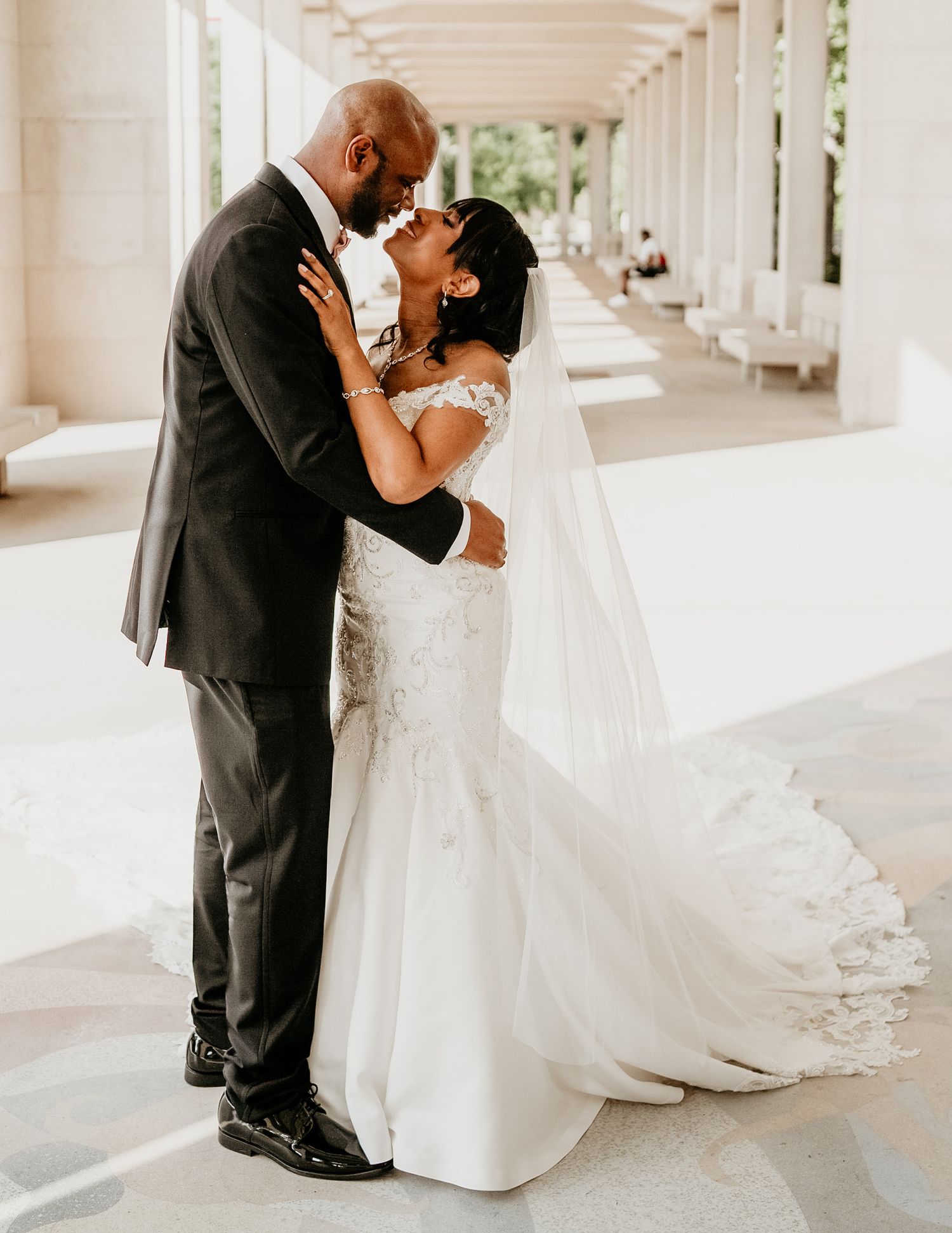 A bride and groom share an intimate kiss in a bright columned corridor, her long white dress trailing behind her.