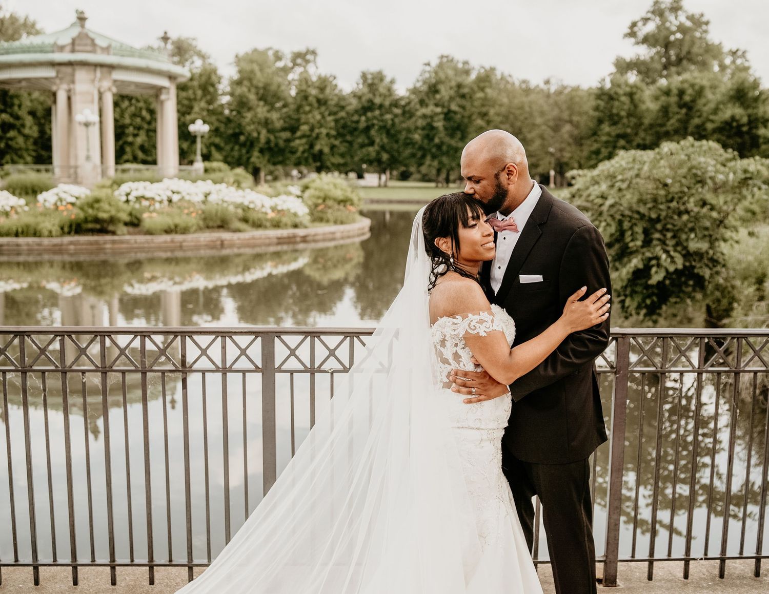A couple shares a romantic moment on an ornate bridge overlooking a pond with a white gazebo in the background.