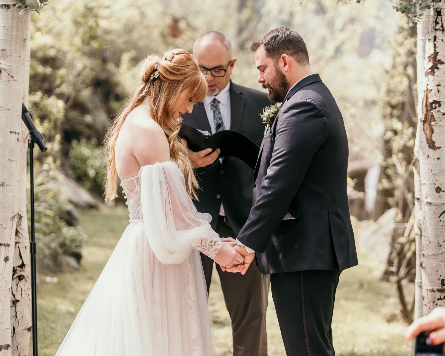 An intimate outdoor wedding ceremony takes place between birch trees with an officiant presiding over the couple.