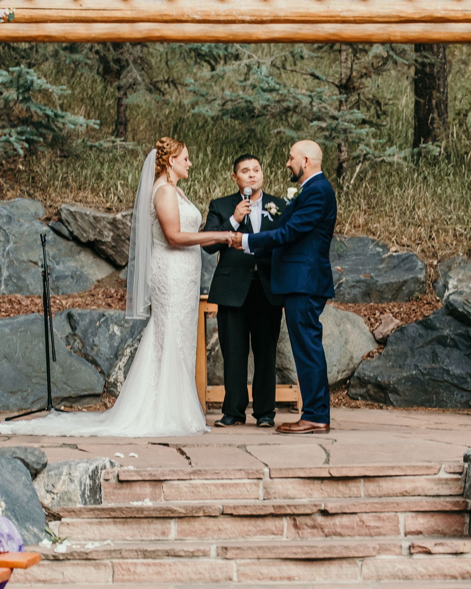 An outdoor wedding ceremony takes place on a stone patio surrounded by natural rock formations and autumn foliage.