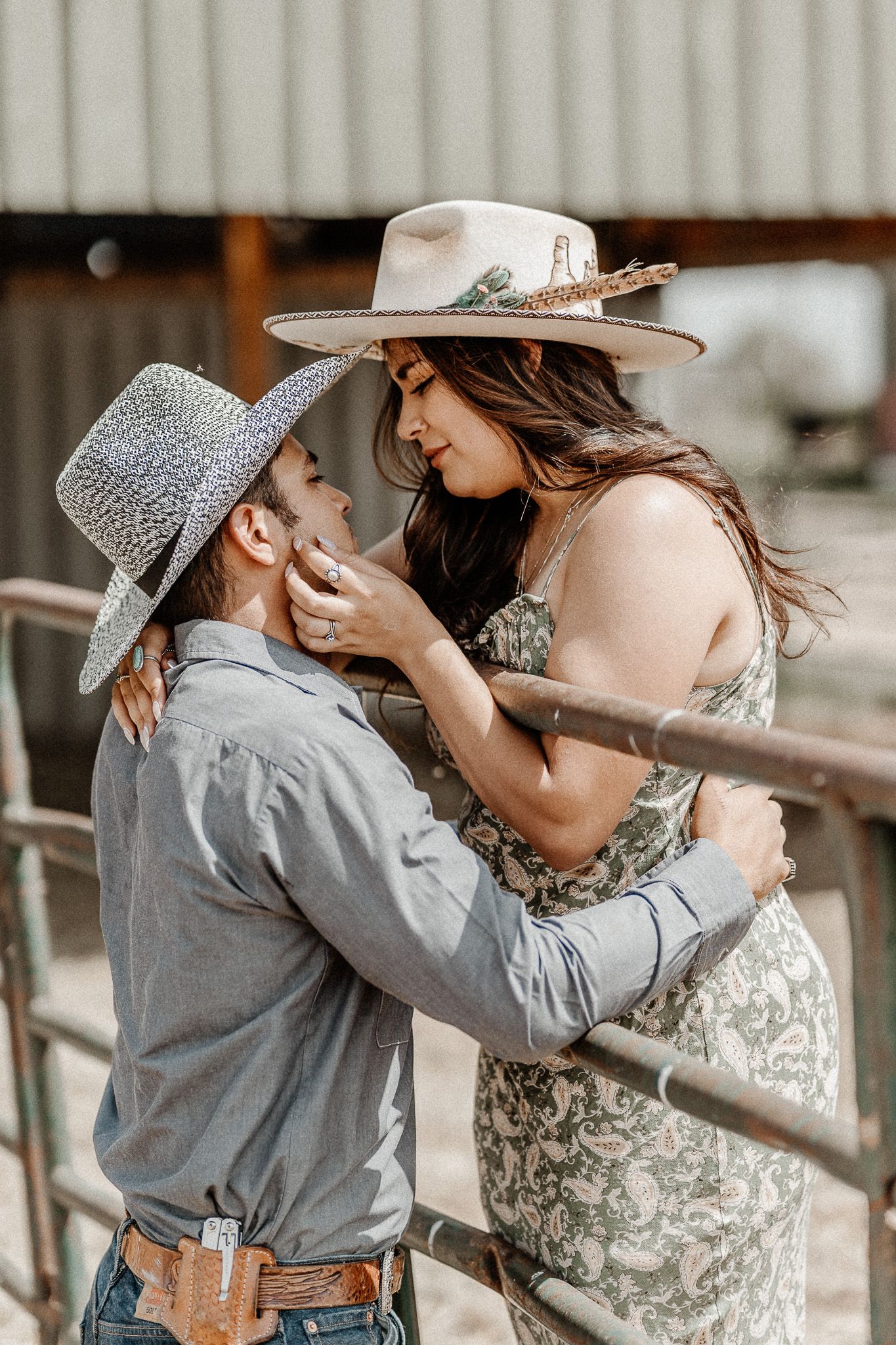 A romantic western-styled couple in cowboy hats share an intimate moment while leaning against a rustic fence.