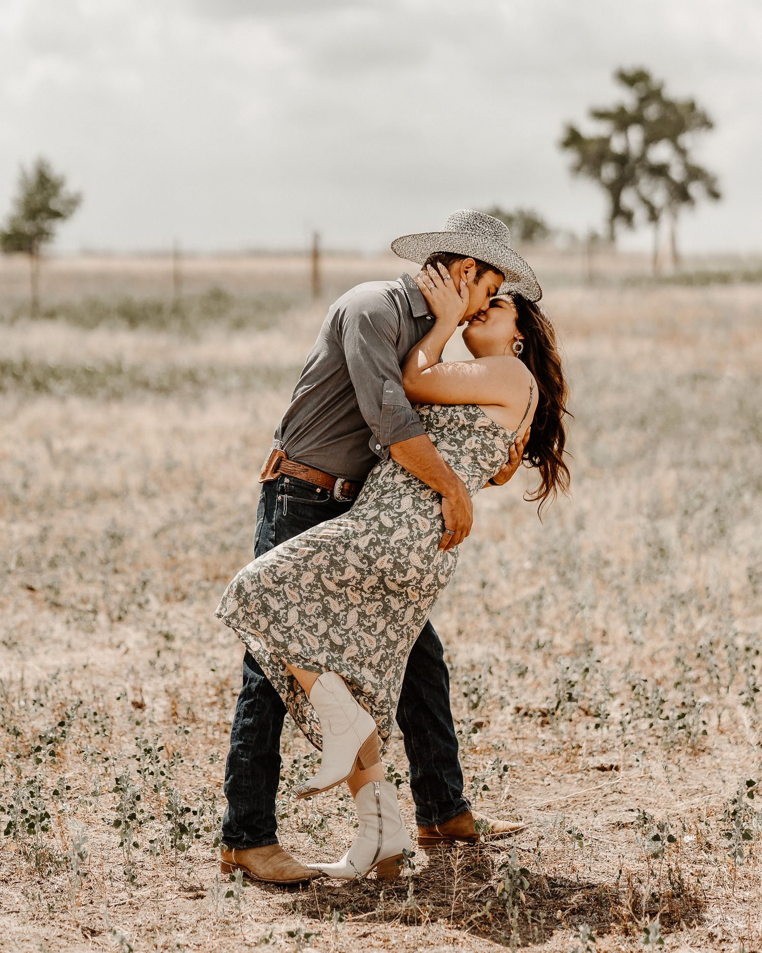 A romantic couple dressed in western style embracing in a sunny deserted field with a palm tree in the background.