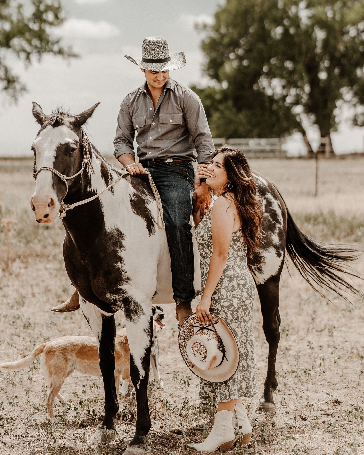 A western-themed couple poses with their horse and dog in a rustic field setting with muted earth tones.