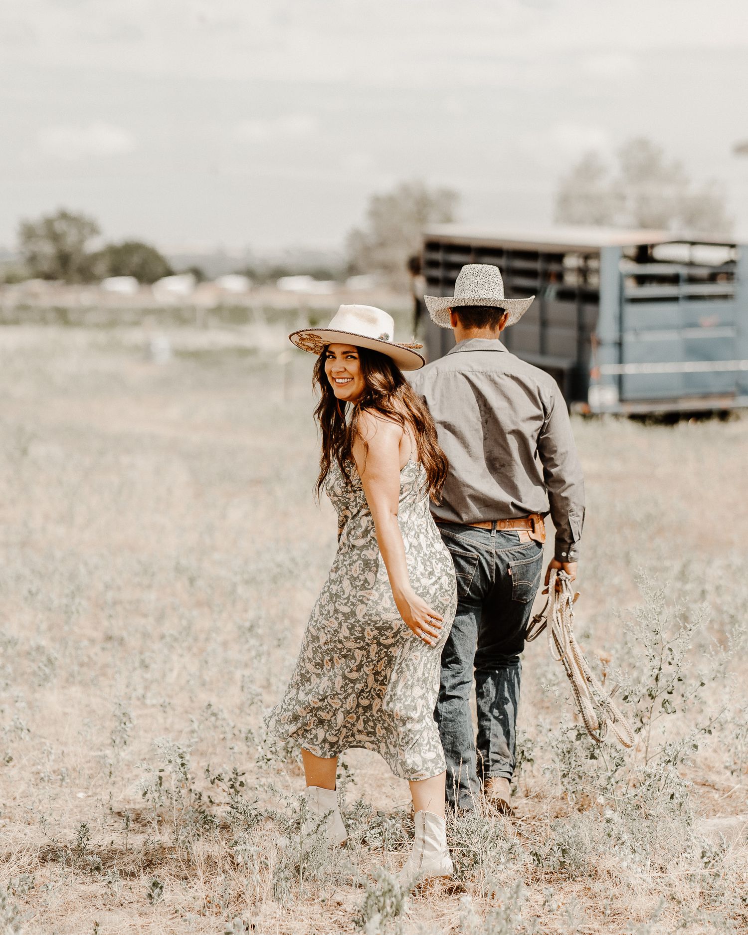 A playful couple in western attire walking through a rural field.