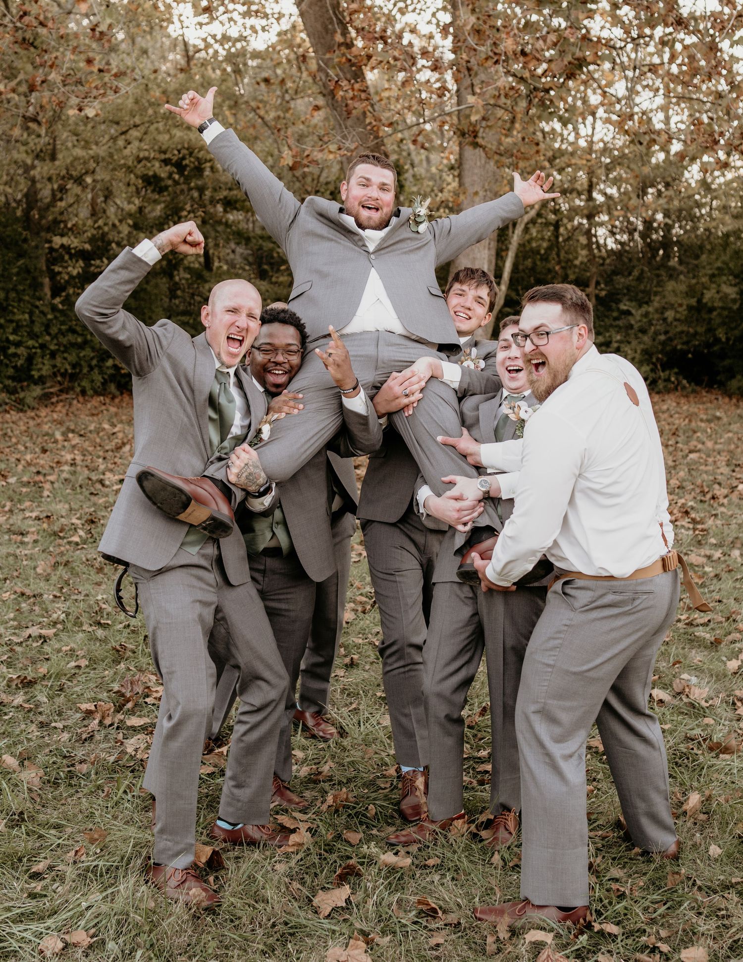 Joyful wedding party in gray suits celebrating and lifting groom in autumn setting at outdoor celebration.