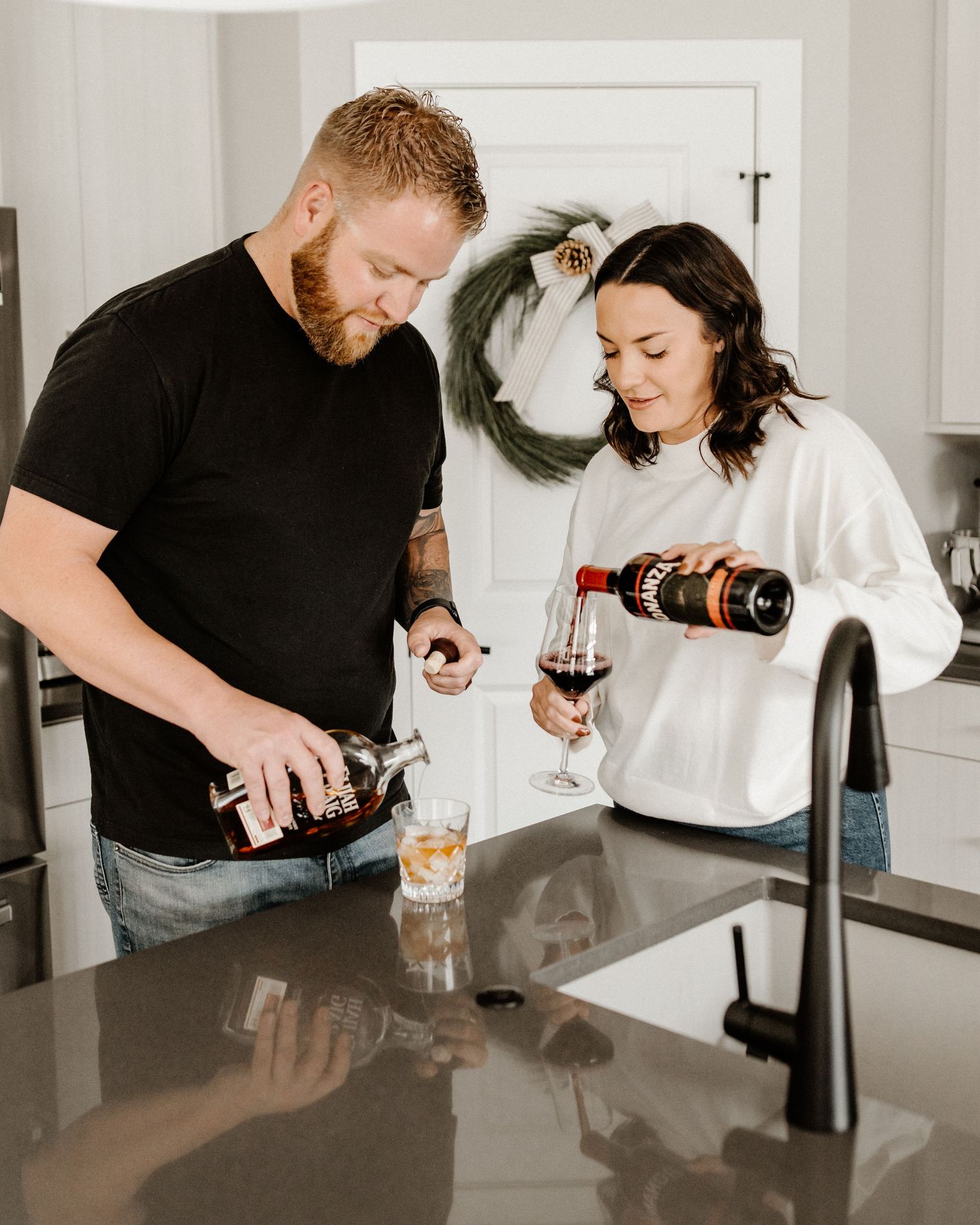 Couple pouring drinks together in a modern kitchen with black countertops and a wreath visible on white door in background.