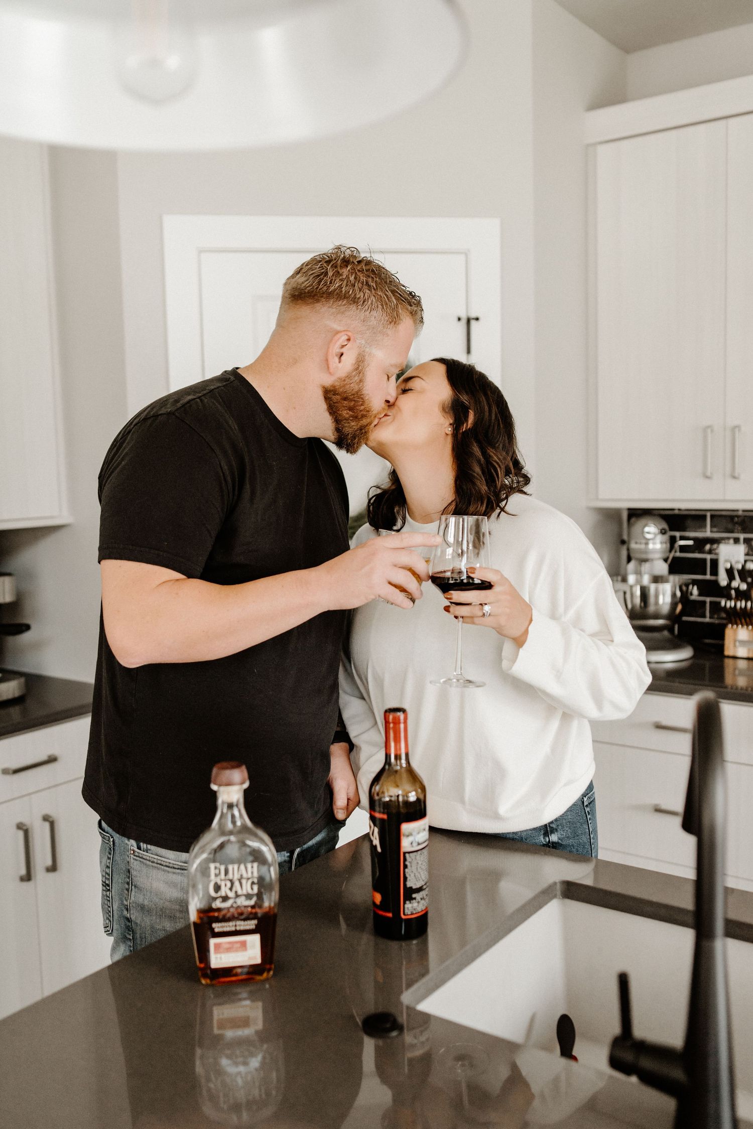 Couple sharing wine and an intimate moment together in a modern white kitchen with bottles on counter.