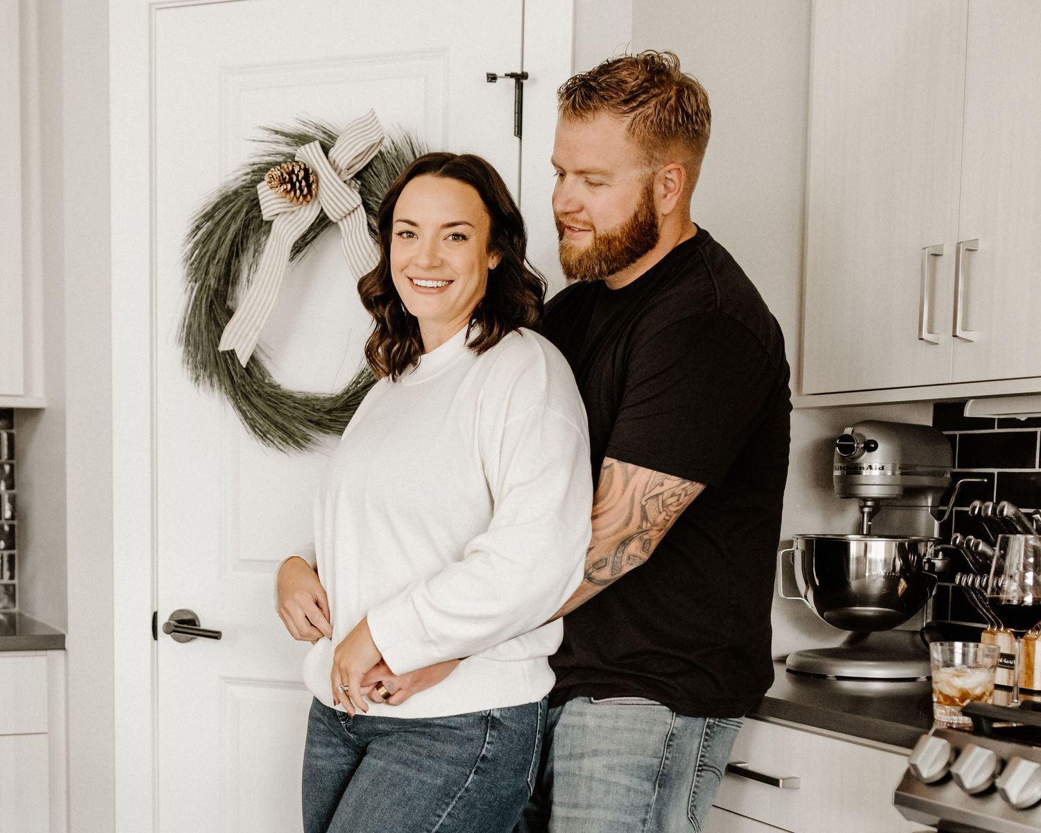 A couple wearing casual sweaters embrace while standing in a bright kitchen with a wreath on the wall.