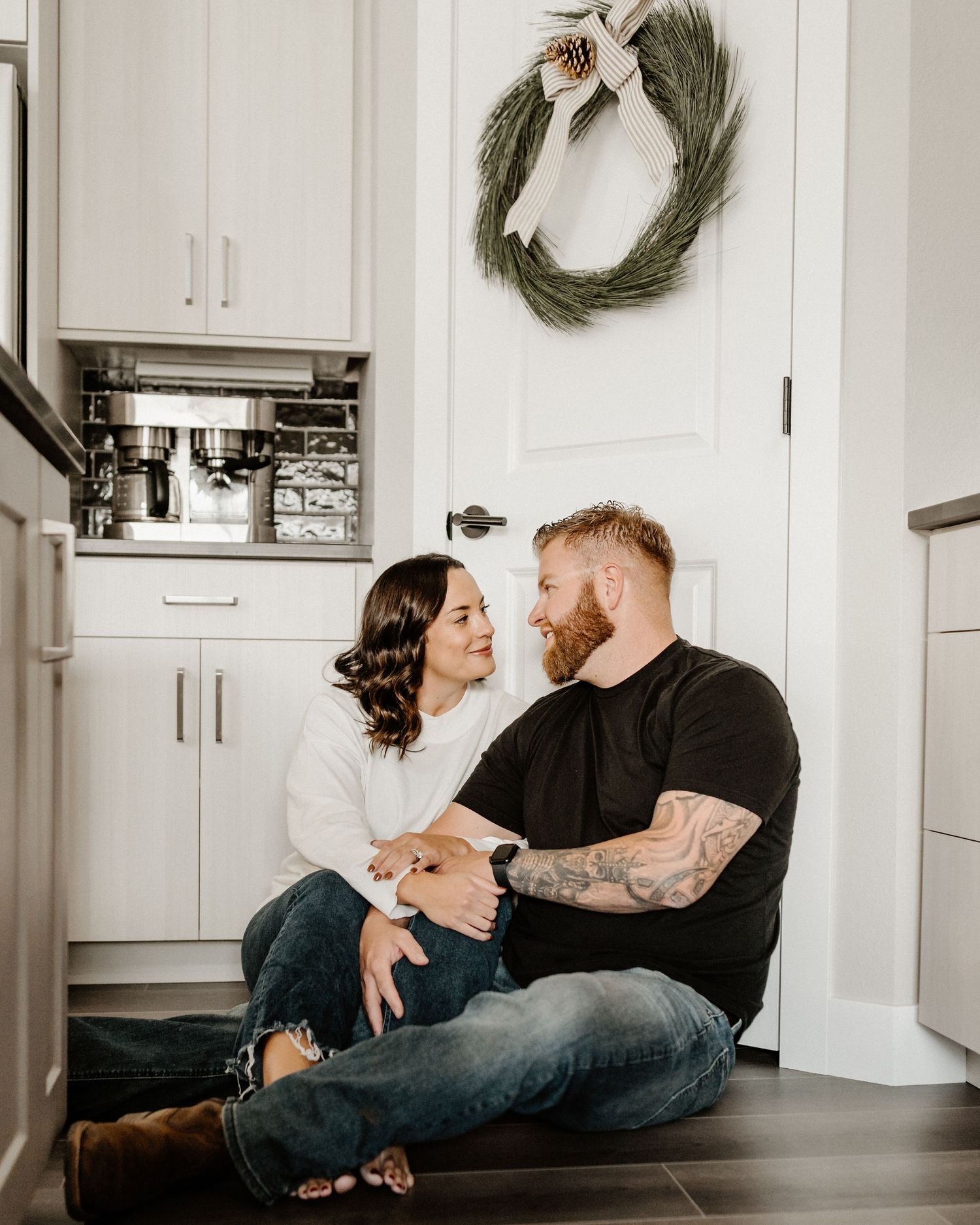 Couple sits together on kitchen floor with wreath hanging on white door in modern farmhouse style interior.
