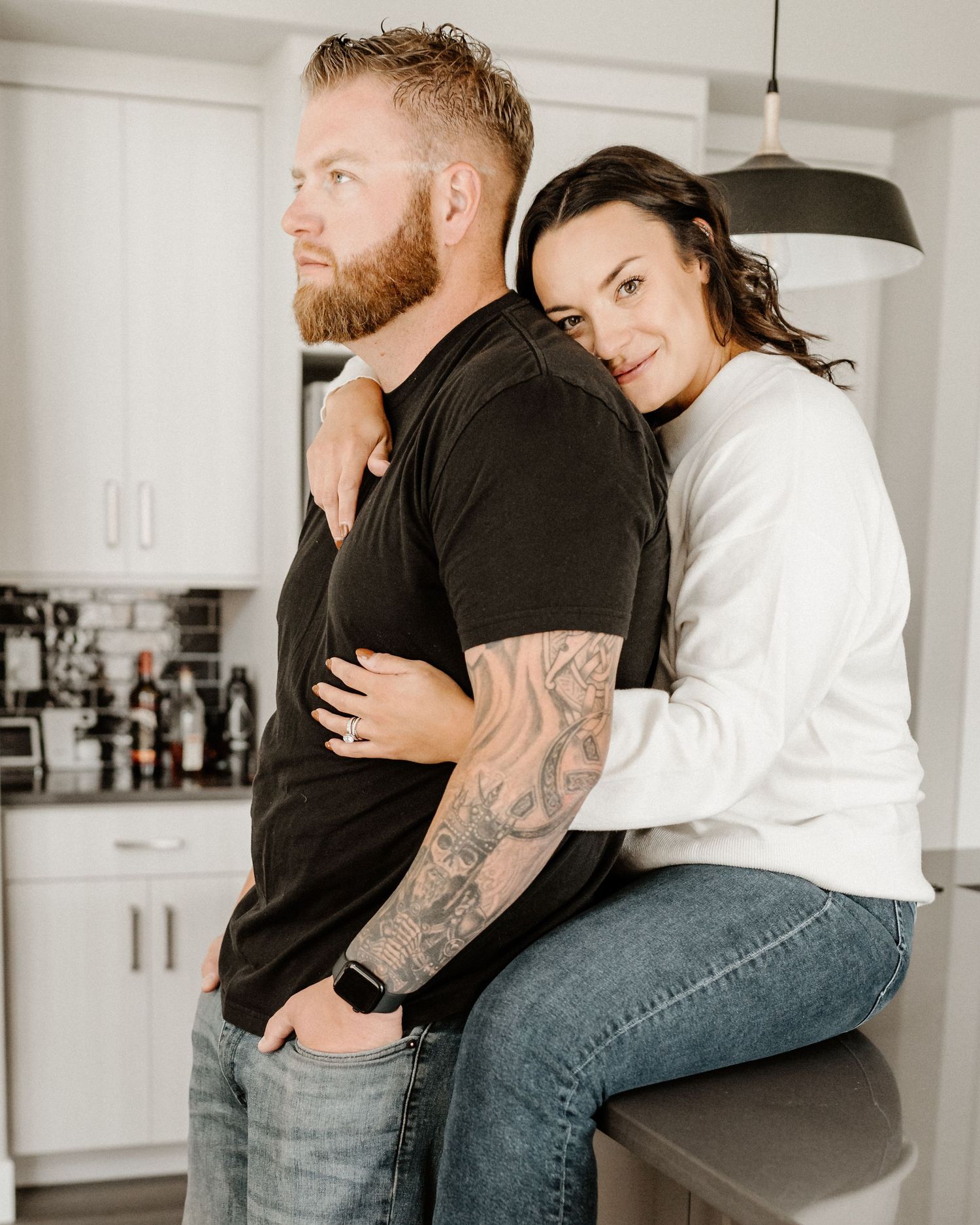 Couple embraces in modern white kitchen with hanging pendant light as woman in white sweater leans on tattooed man in black.