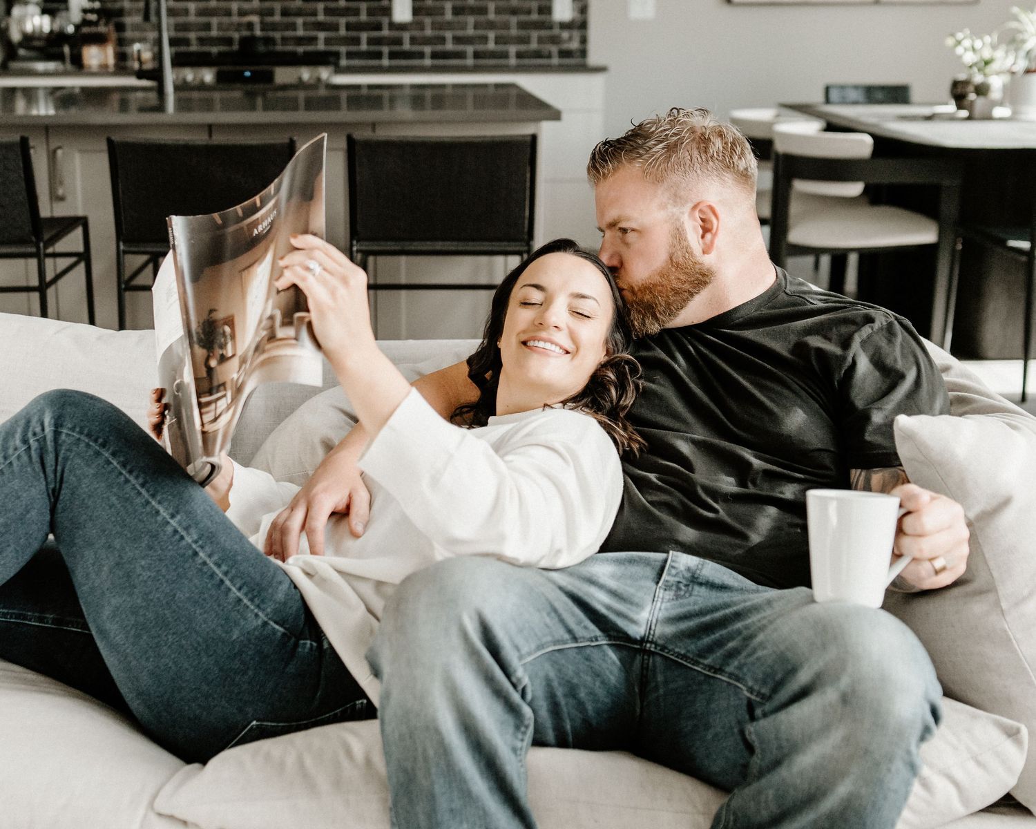 Couple relaxes together on gray couch at home with coffee and newspaper in a cozy living room setting.