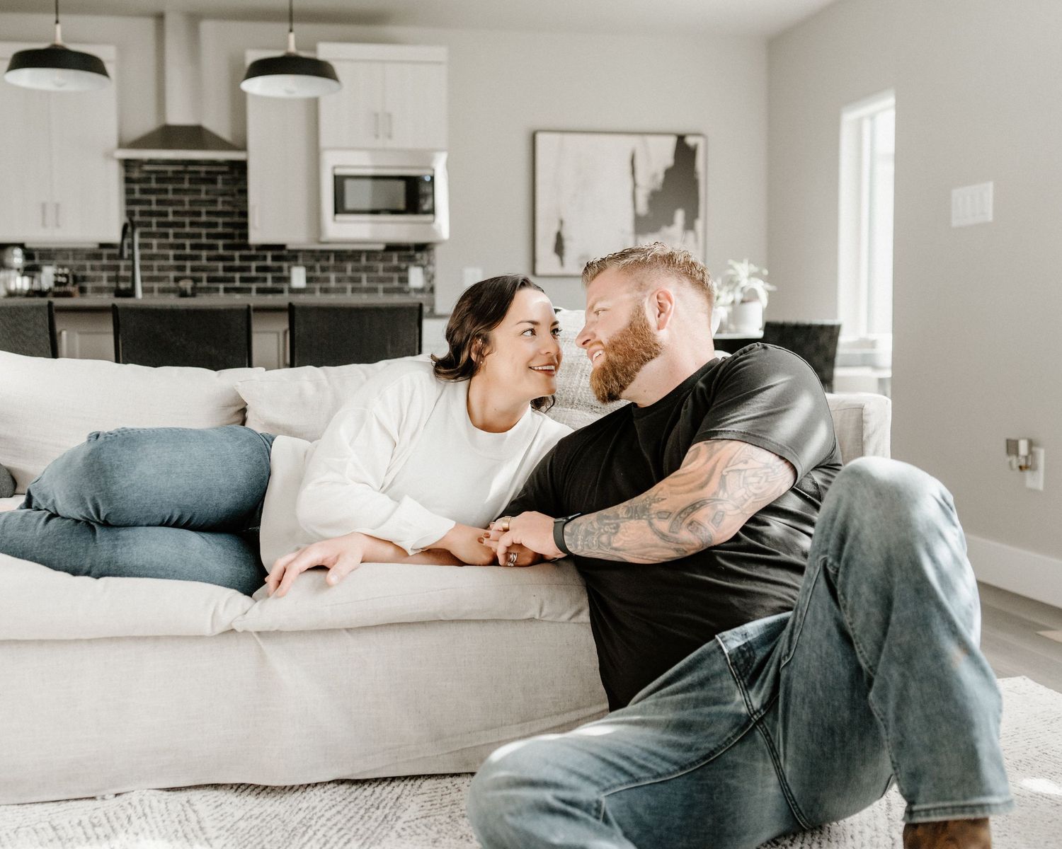 A couple relaxes together on a light gray couch in a modern living room with black pendant lights and brick accents.