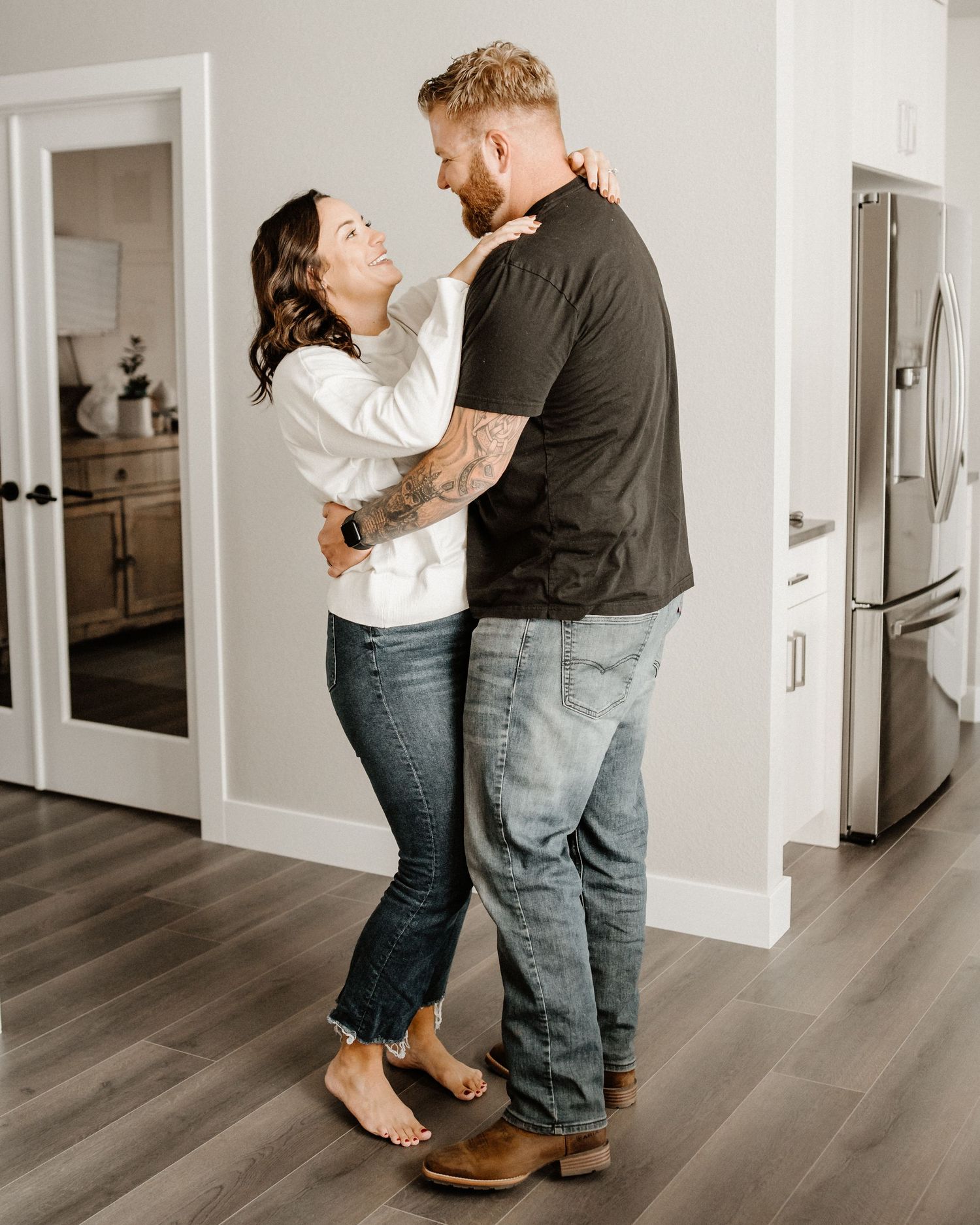 A couple share an intimate embrace while dancing in their kitchen with dark hardwood floors and white walls.