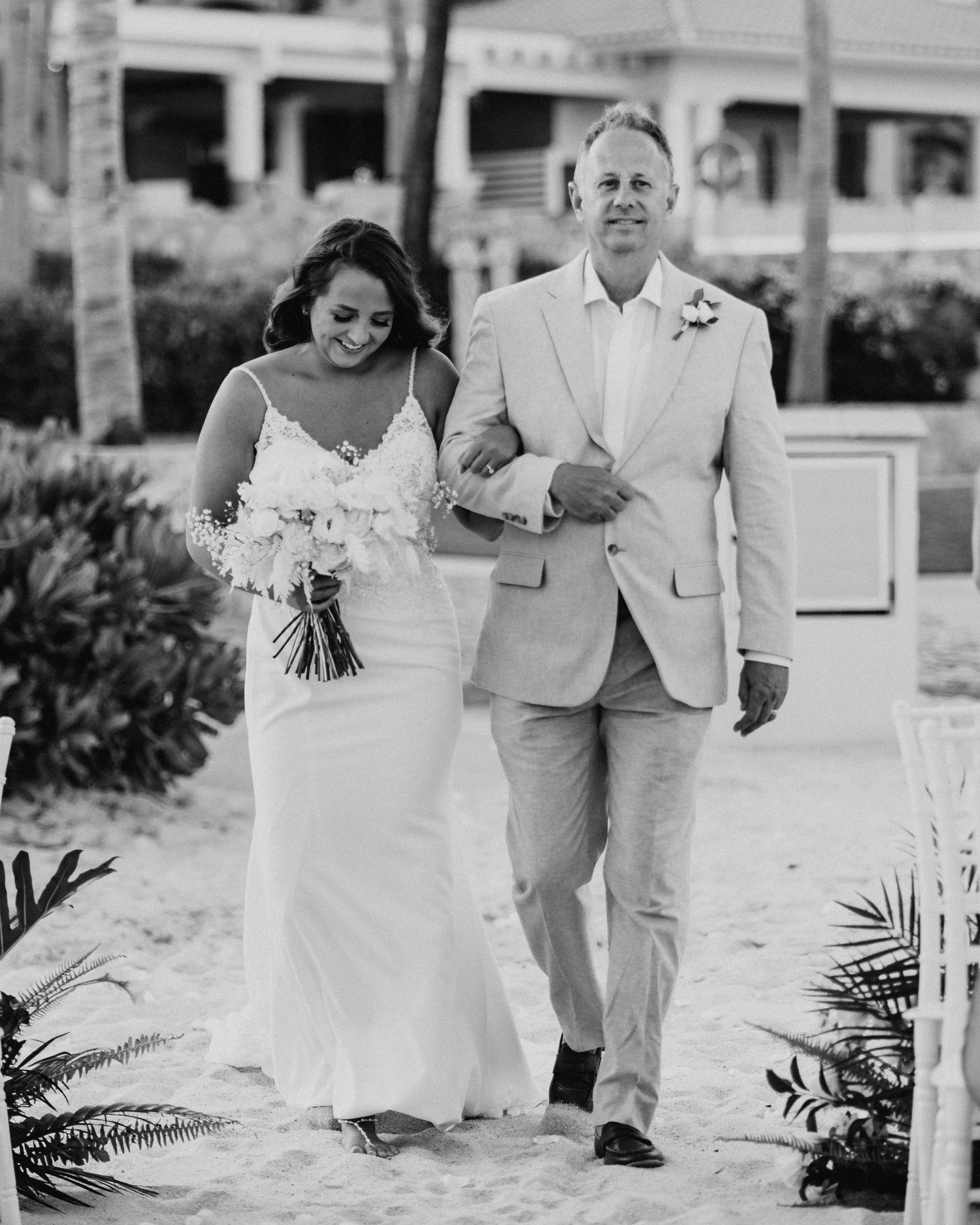 A wedding couple walk arm in arm down a palm-lined path in black and white, the bride holding a white bouquet destination wedding Cabo