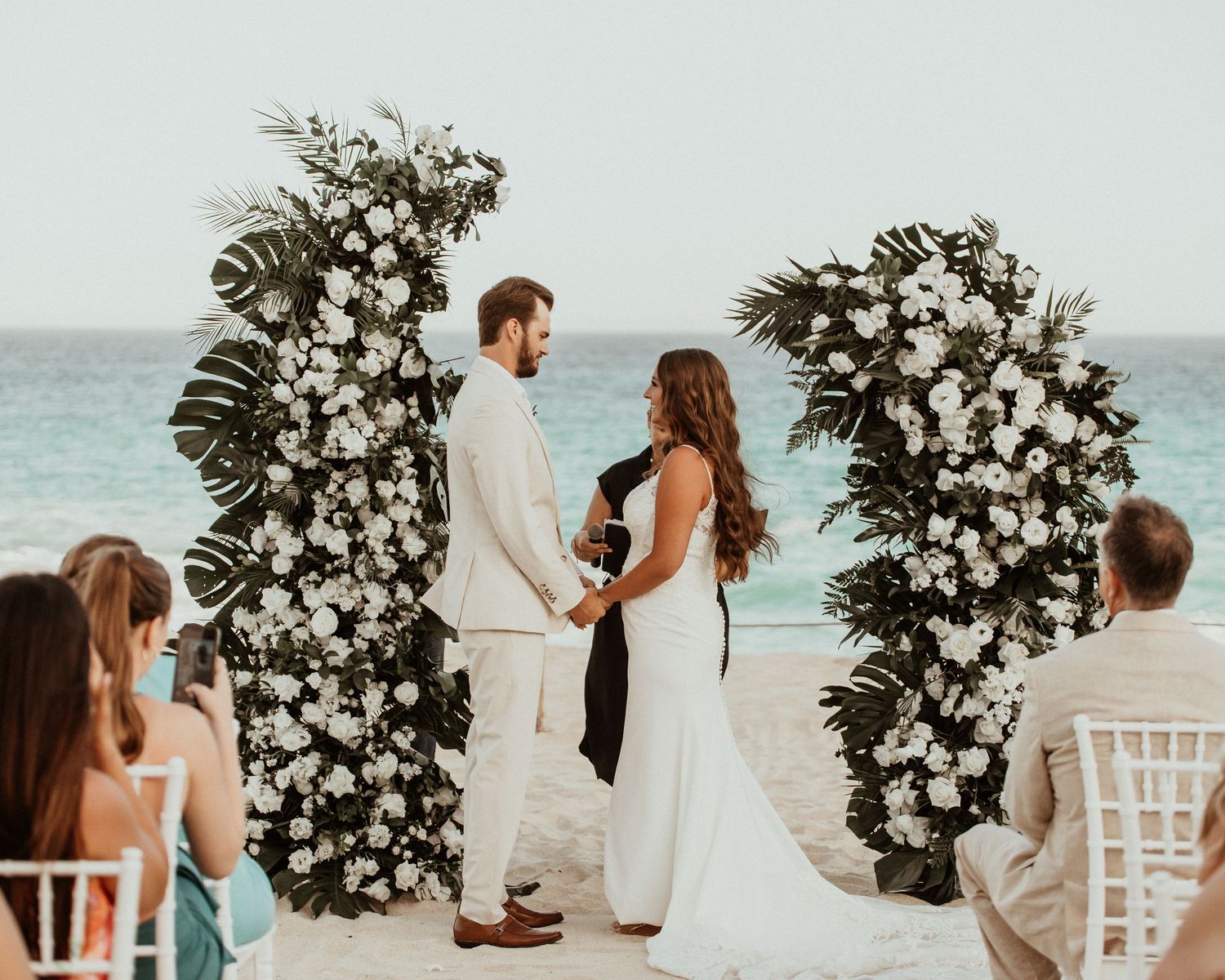 A beachfront wedding ceremony with two people standing between white floral arches on the sand by the ocean. destination Cabo San Lucas