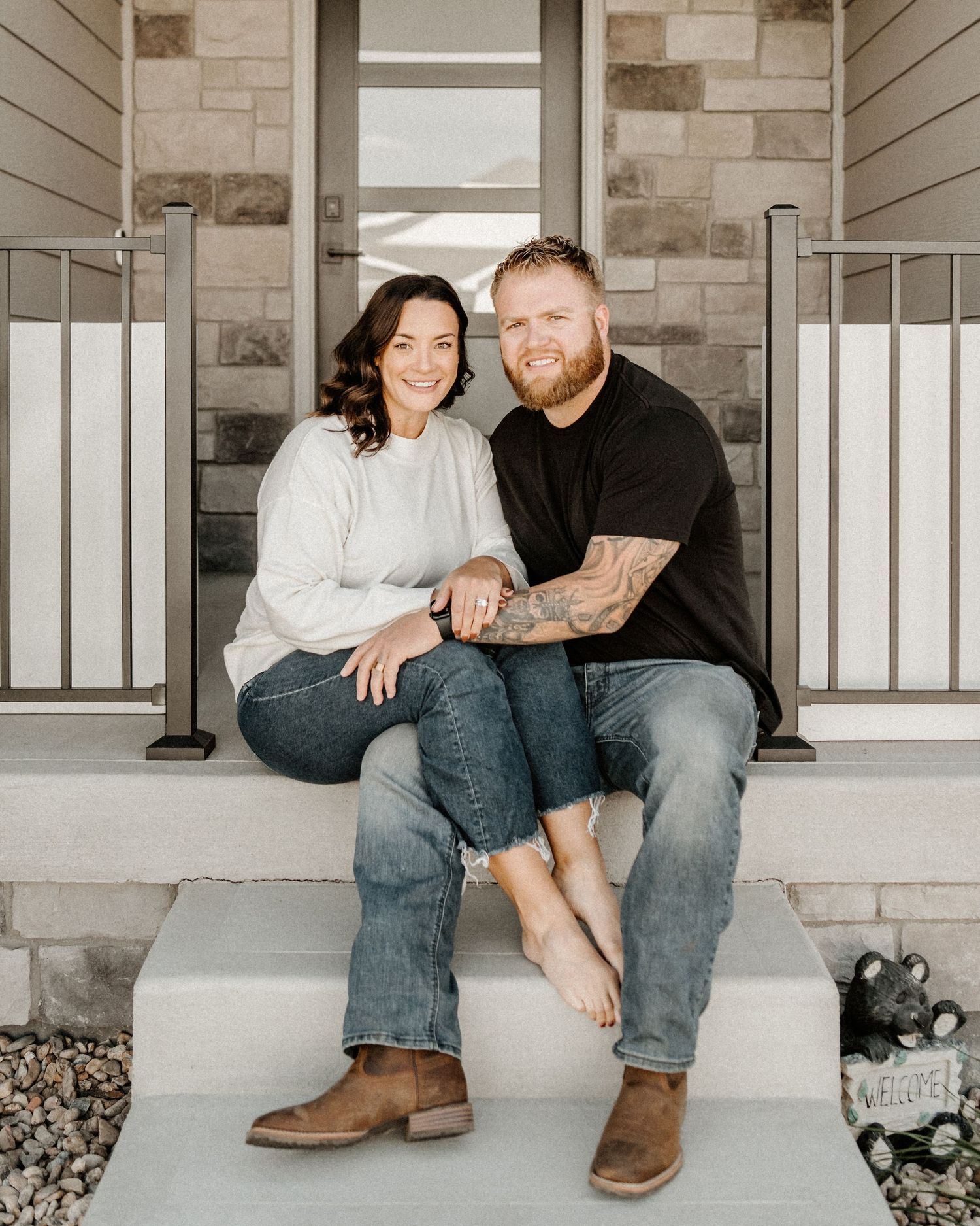 A couple sits together on concrete front porch steps of stone and siding house wearing casual clothes.