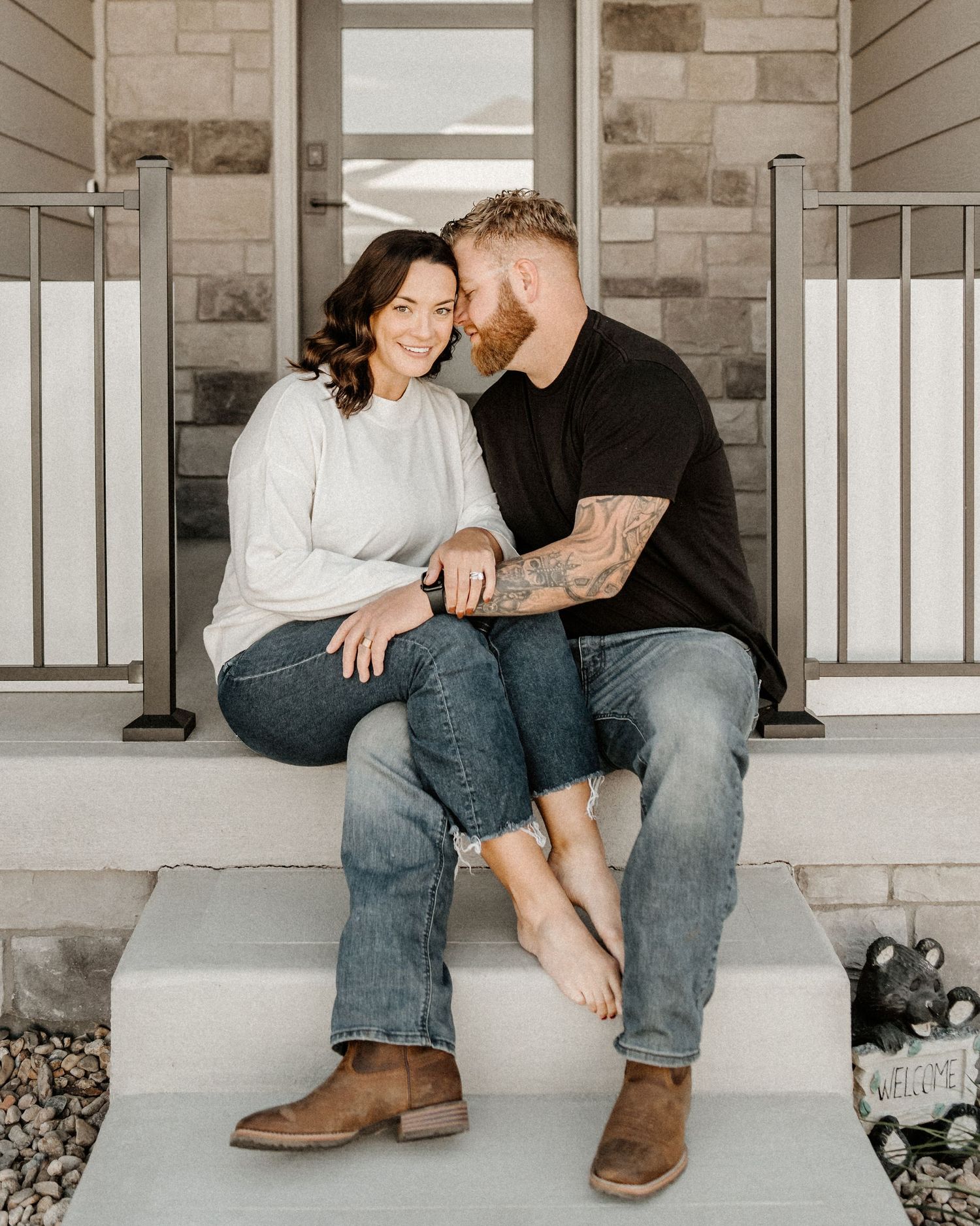 Couple sits close together on front porch steps of a stone and siding house wearing jeans and boots.