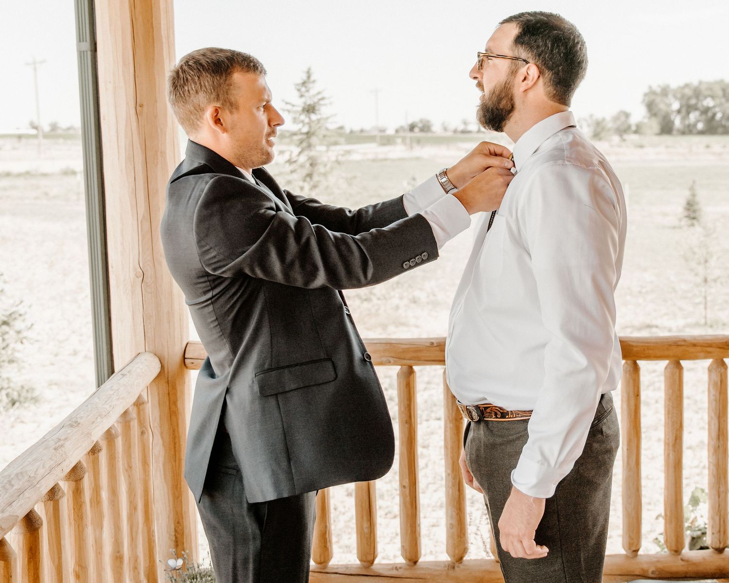 Best man helps adjust groom's tie while standing on a wooden porch during wedding preparations on a sunny day.