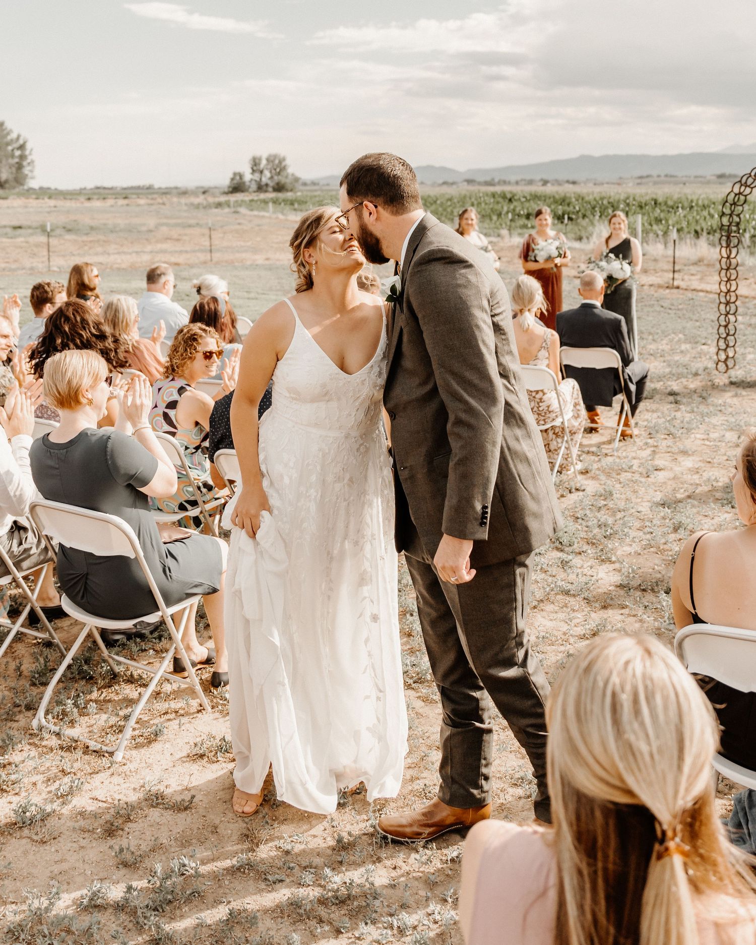Newlyweds share an intimate moment during outdoor ceremony at rustic vineyard with guests seated on folding chairs.