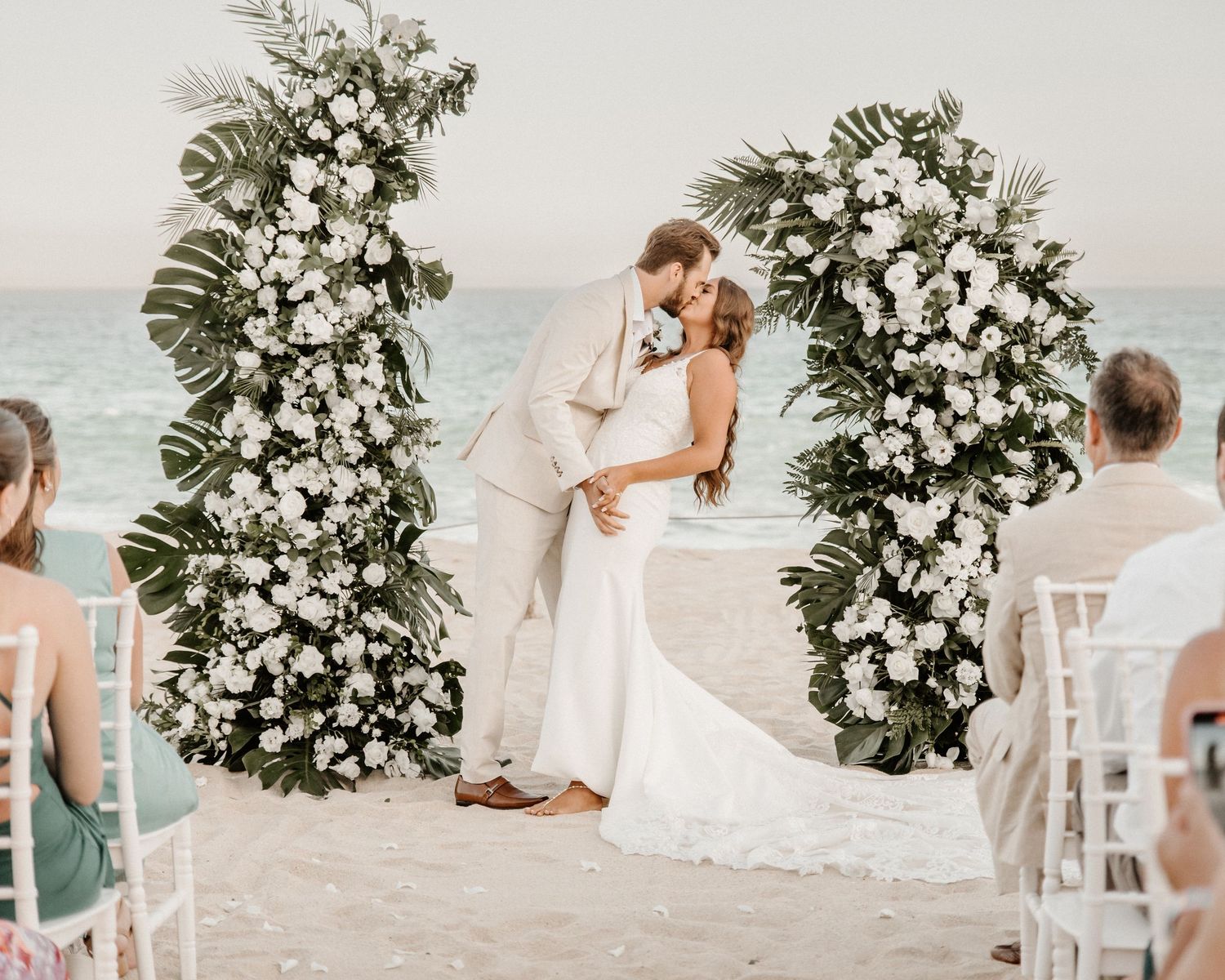 Romantic beach wedding ceremony with white floral archway and palm fronds overlooking the ocean at sunset.
