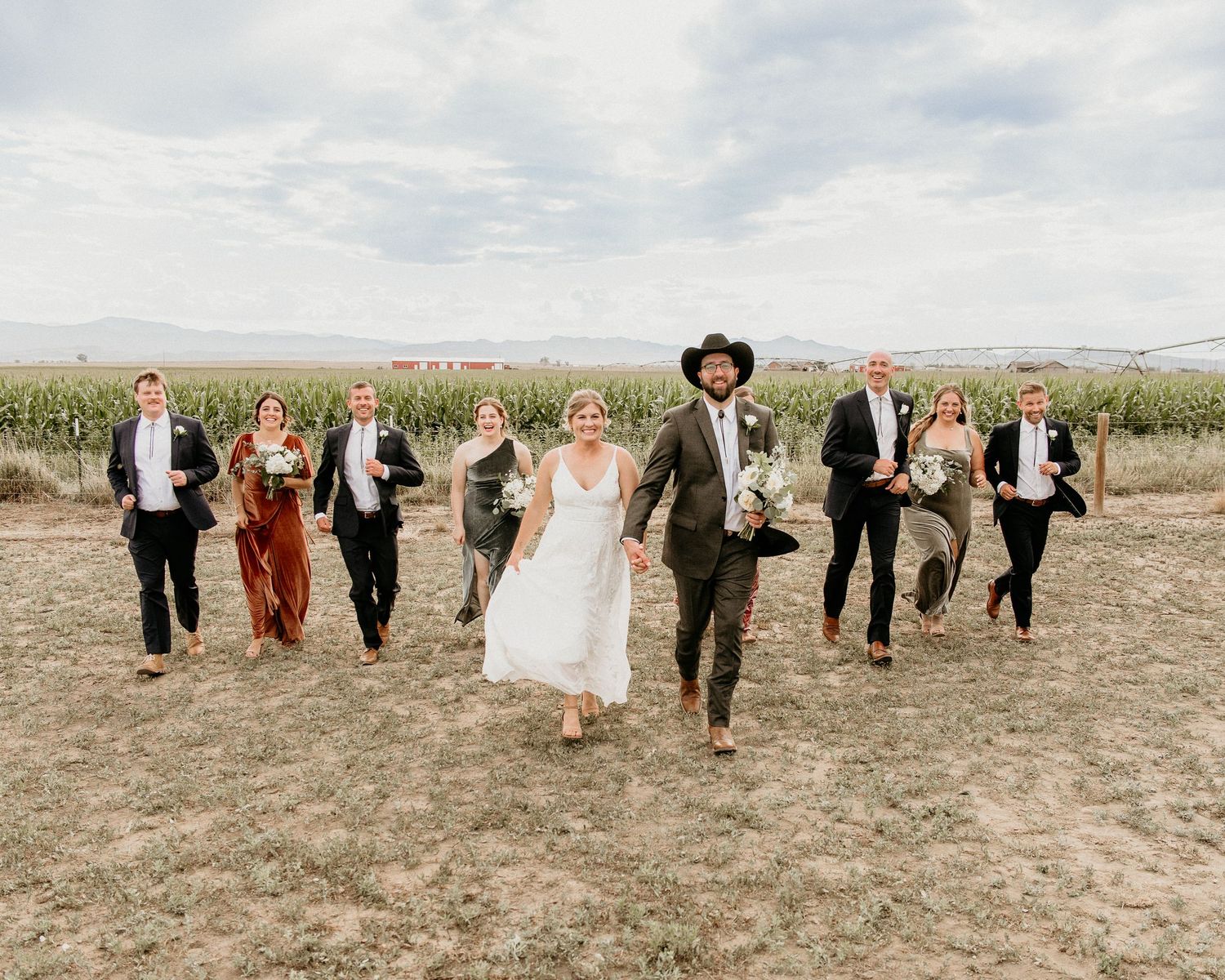 Wedding party runs joyfully through a field with mountains in the background on a cloudy day.